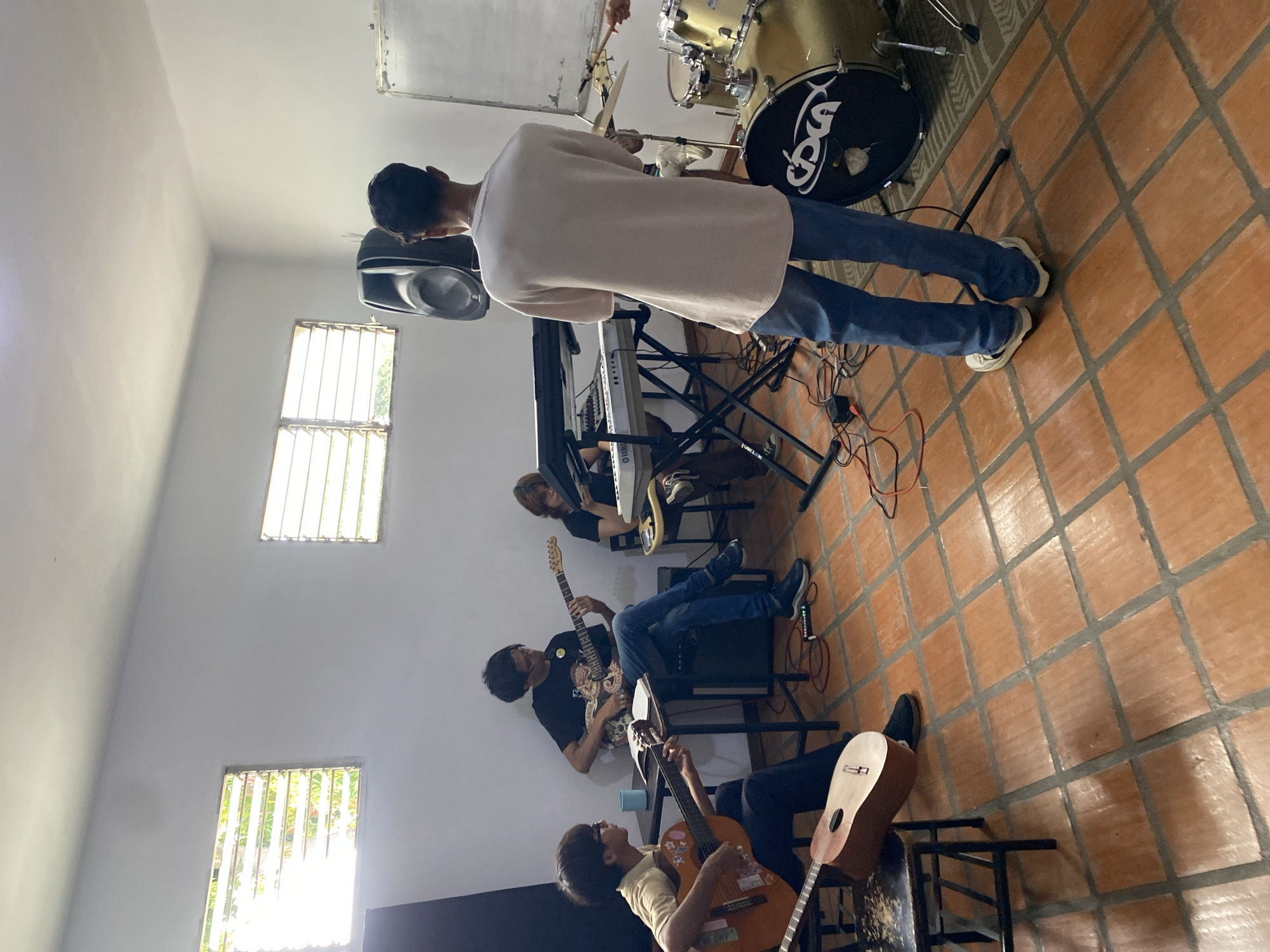 Four young people are in a room, seemingly practicing music. Two are playing acoustic guitars, one is playing an electric guitar, and another is seated at a keyboard. A drum set is visible in the background, along with audio equipment including a speaker. The room has white walls and two windows with horizontal blinds. The floor is tiled with terracotta-colored squares. This appears to be a rehearsal space or a music studio located in Tinaquillo, Venezuela.