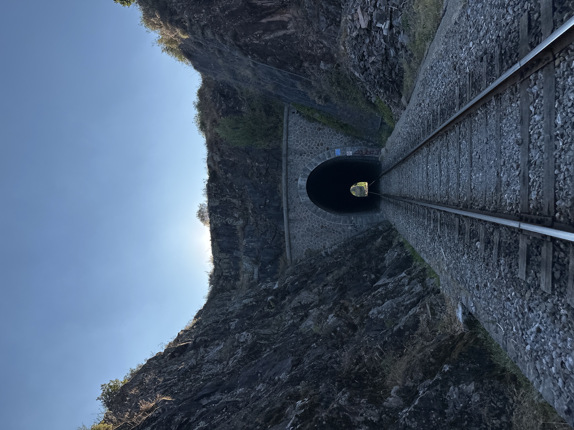 This is a low-angle, wide shot of train tracks leading into a stone train tunnel. The tunnel entrance is arched and constructed from rough-hewn stones, with a smaller, arched opening visible within the larger archway. Light streams from the tunnel's exit, silhouetting the interior. The tracks are made of steel rails and wooden ties, laid on a bed of gravel. On either side of the tracks, steep, rocky hillsides rise sharply. The left hillside is covered in dark, rough rock with sparse vegetation. The right hillside also consists of rock and vegetation, with some areas reinforced with netting. The sky above is clear and a pale blue, with the sun low on the horizon, casting a warm glow. The overall environment suggests a remote, natural setting. This location is the Tunnel de Générargues, located in Générargues, France.