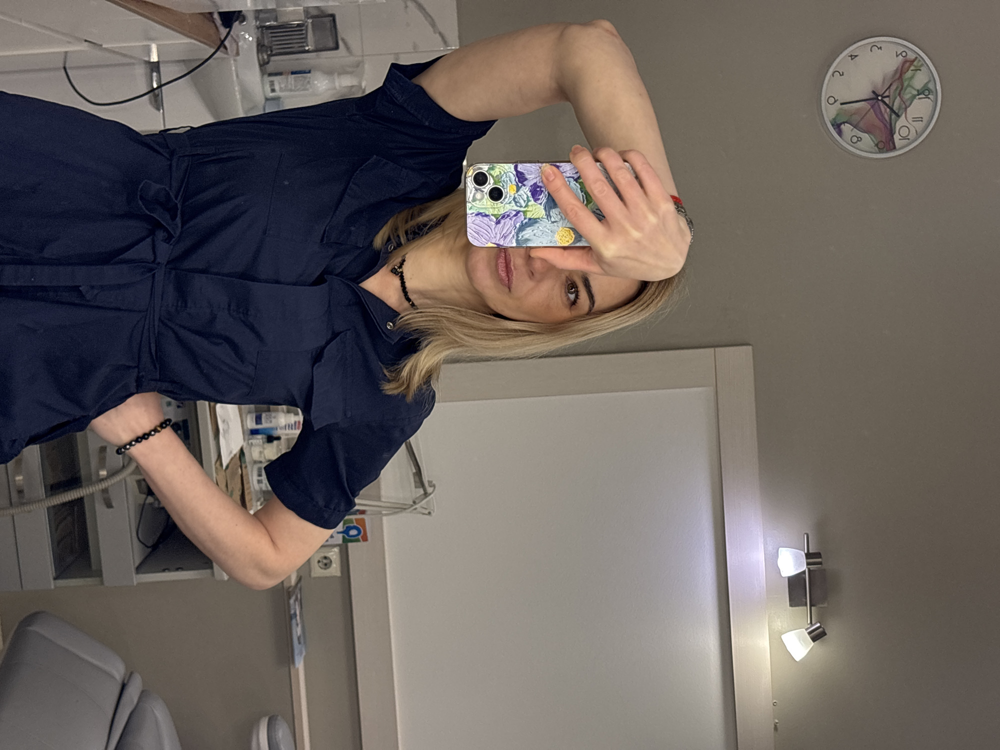 A woman is taking a selfie in a room, likely a medical or dental office, judging by the equipment visible. She's wearing a navy blue dress and has blonde hair. Her right hand holds a phone with a colorful case in front of her face. She's partially blocking her face with the phone. On the wall behind her, there's a clock showing approximately 1:55. A white doorframe and a wall-mounted light fixture are also visible. The location is Unknown, Unknown.
