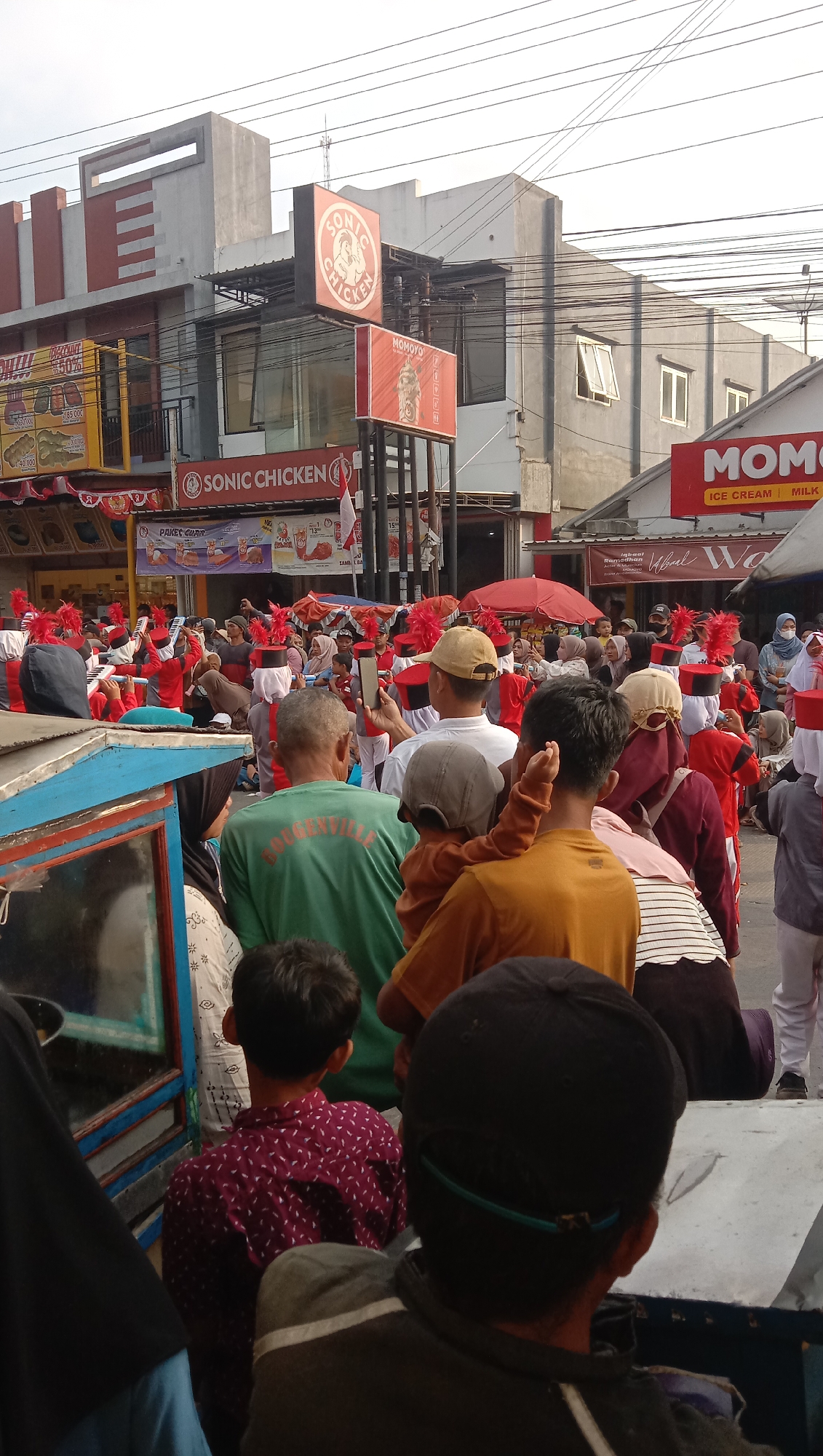 This photograph, taken in the city of Jatisawit, Indonesia, captures a lively street scene, likely during a parade or celebration. A group of people in the foreground, facing away from the camera, are watching an event unfolding in the distance. Among them, a child is being held by an adult, both looking towards the commotion.
The main focus of the image is a procession of individuals dressed in red uniforms, adorned with red feather headdresses and black hats with red bands. They appear to be marching or performing, with some holding instruments or flags. The crowd surrounding them is diverse, including men, women, and children, many of whom are observing the performance and some are capturing it with their mobile phones.
Behind the procession, commercial buildings line the street. Signs for "SONIC CHICKEN" and "MOMOYO" are prominently visible, indicating food establishments. Indonesian flags are also displayed, suggesting a national celebration or event. The sky above is overcast, with power lines crisscrossing overhead, a common sight in urban Indonesian settings. The overall atmosphere is one of community engagement and festive spirit.