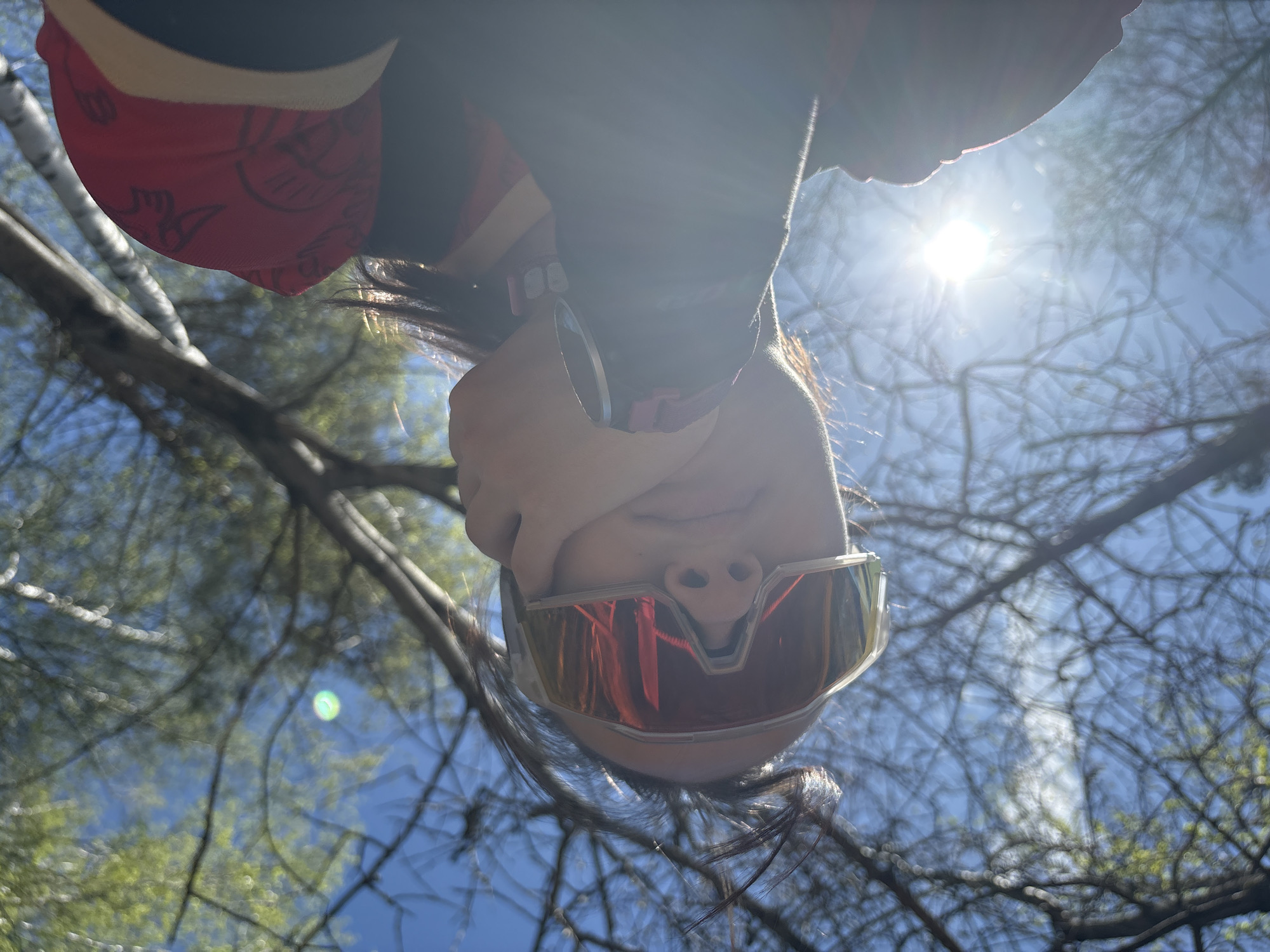 The image is an upside-down, low-angle shot of a person wearing mirrored sunglasses and a red head covering with a pattern, set against a backdrop of branches and a bright sky with the sun visible. The person's face is partially obscured by the sunglasses and the angle of the shot. The background reveals tree branches against a blue sky with a bright sun. The scene is outdoors. The image cannot be located at the city of Perm, Russia based on its visual content.