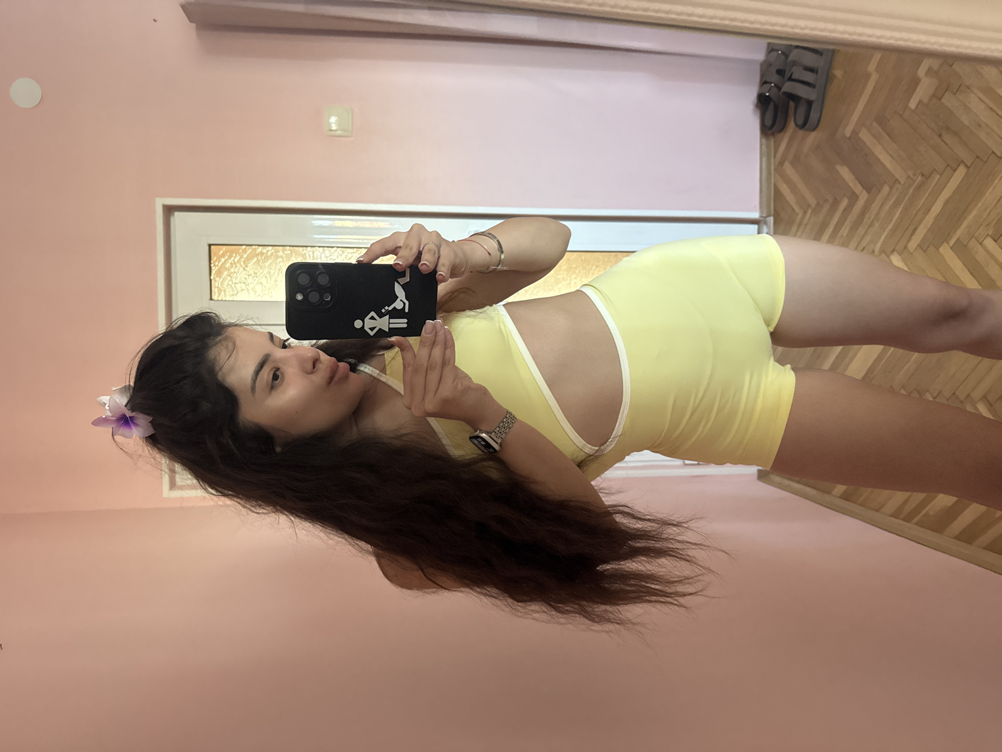 A woman with long, dark hair is taking a selfie in what appears to be an indoor setting. She is wearing a light yellow two-piece outfit consisting of a top and shorts. A flower is visible in her hair. She is holding a black smartphone with a design on the back case. The background features a pink wall, a white-framed door with frosted glass, and a wooden floor. Two pairs of sandals are visible near the top edge of the frame. It is not possible to pinpoint the exact location within Plovdiv, Bulgaria without additional information.