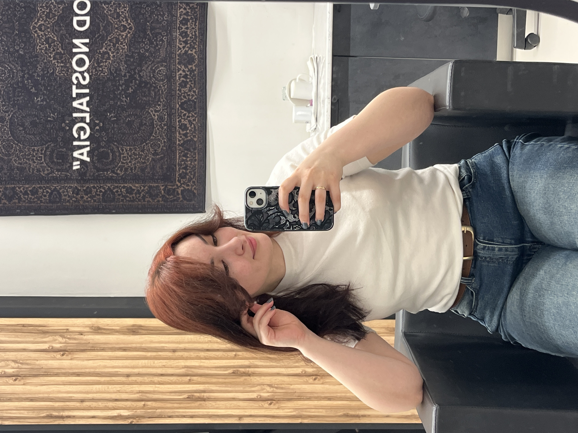 A young woman with auburn hair is taking a selfie in a mirror. She is sitting in a black chair with a brown belt around her jeans. Behind her is a wall with a rug hanging on it, and a wooden surface beneath. The rug is dark with lighter-colored patterns and the text "OFF NOSTALGIA CO." There is no visible evidence to suggest that the location is Neftekamsk, Russia.
