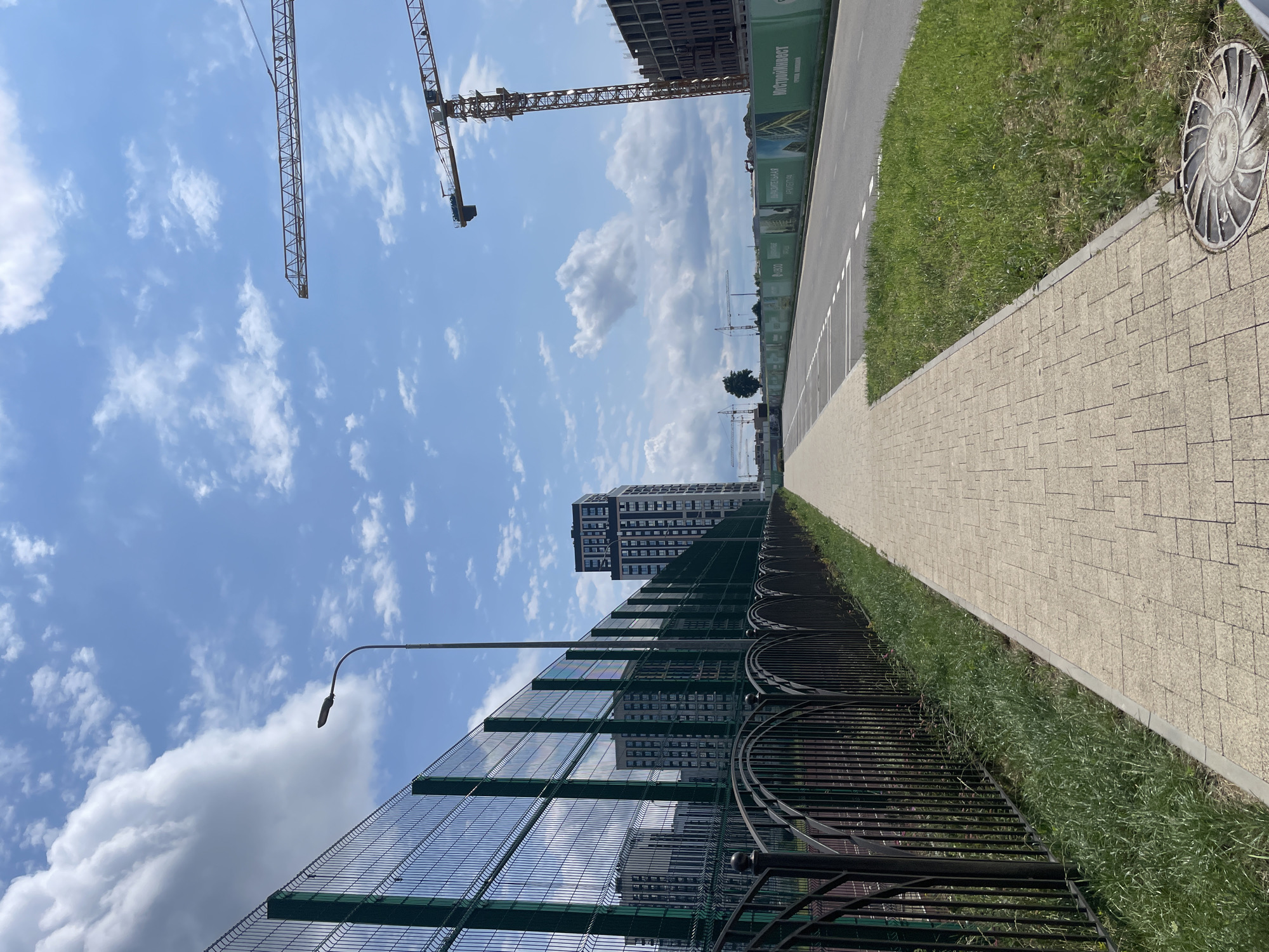 The image depicts a construction site in Stavropol, Russia, under a bright, partly cloudy sky. A paved sidewalk runs alongside a black metal fence, which in turn borders a green wire mesh fence. Beyond the fences, several multi-story apartment buildings are visible, some completed and others under construction. Towering yellow construction cranes dominate the skyline, indicating ongoing development. A road with white lane markings runs parallel to the sidewalk and construction area, and a manhole cover is present on the grassy verge. The scene is devoid of people.