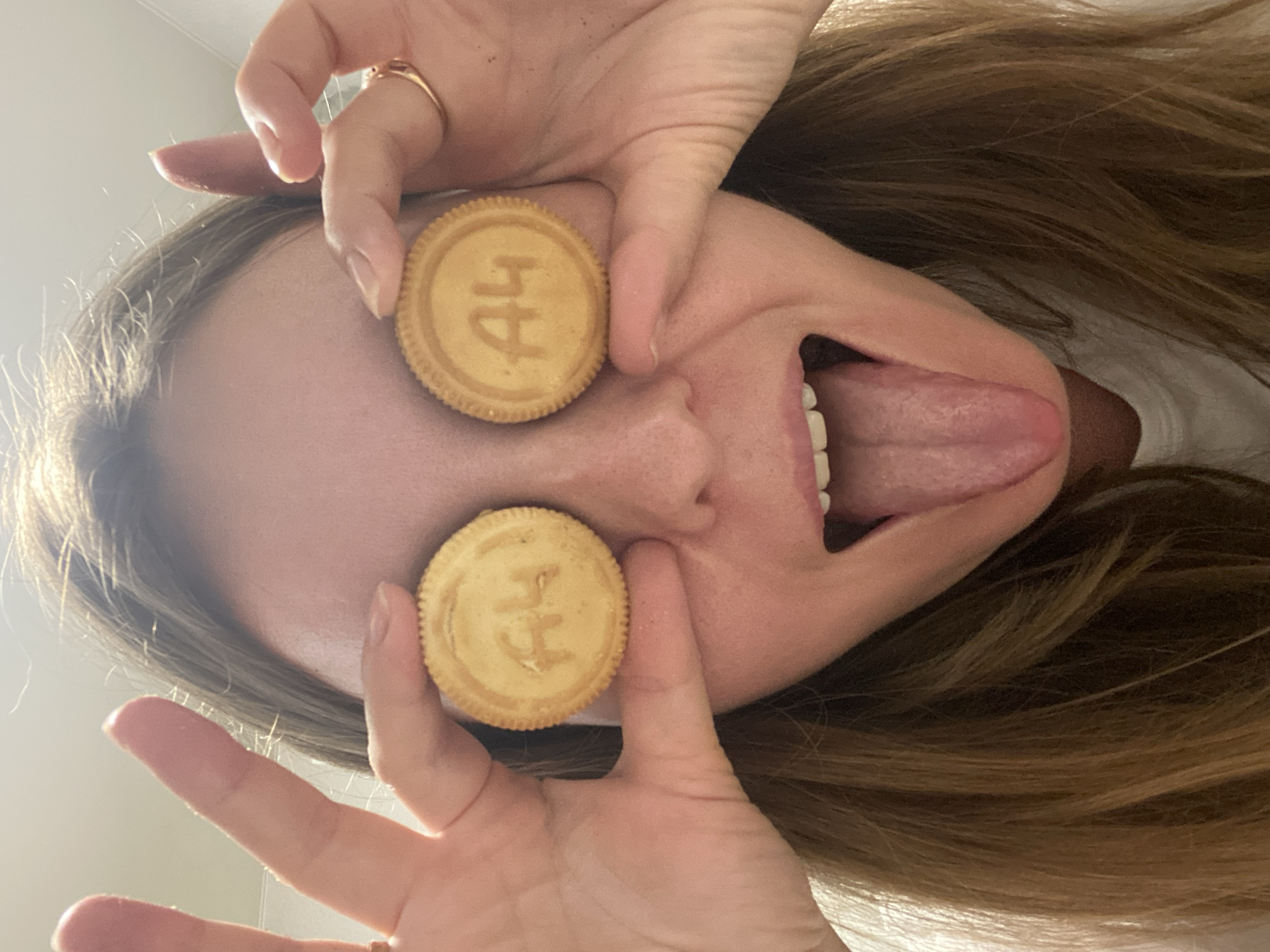 This low-angle, close-up image, taken in Noviy, Russia, captures a person's upside-down face making a humorous expression. The subject, likely a young adult with long, light brown hair, is holding two round, light-colored biscuits over their eyes with both hands. Their mouth is wide open, and their tongue is playfully sticking out.
On the biscuit covering the person's left eye (viewer's right), the text "A4" is clearly visible. The biscuit covering the person's right eye (viewer's left) features a symbol that resembles an anchor or stylized letters "HH". Both biscuits have distinct ridged edges. A ring is visible on the person's right hand. The background is a brightly lit, out-of-focus, plain light surface, suggesting an indoor setting with ample daytime illumination. The image conveys a lighthearted, silly, and playful moment.