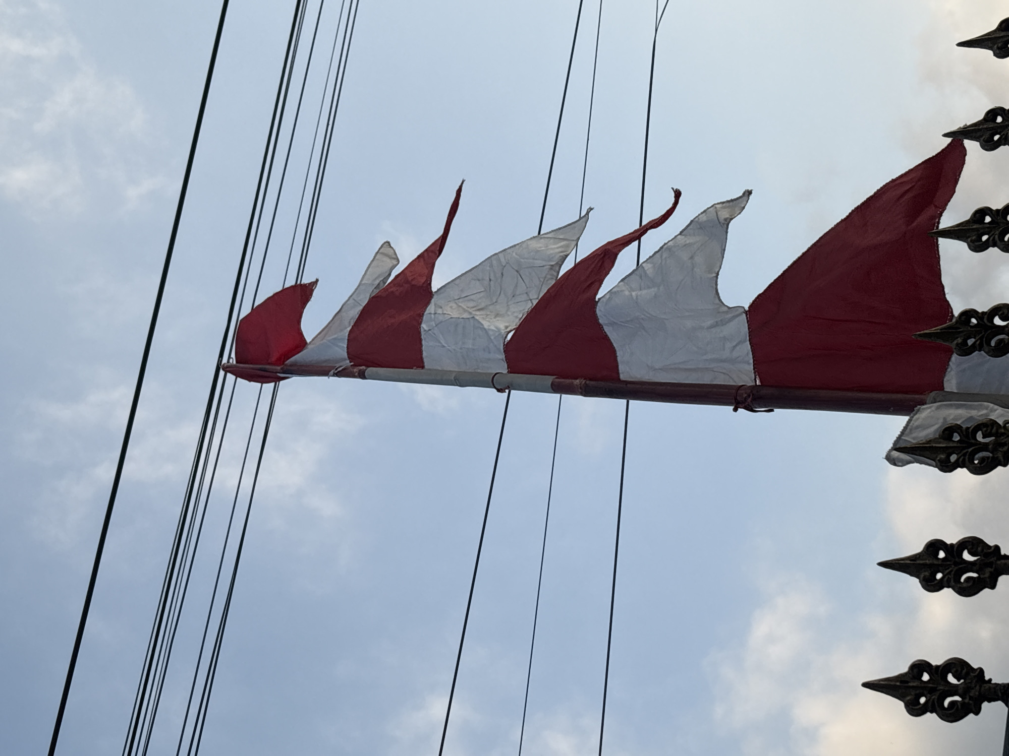 The image shows a close-up of a flagpole with several red and white pennant flags, possibly representing the Indonesian flag. The flags are tattered and appear to be old. They are attached to a pole that extends upwards. Behind the pole and flags, several power lines are visible against a cloudy blue sky. In the foreground, the decorative tops of a dark metal fence are visible. The scene appears to be outdoors, and the perspective suggests the photo was taken from a low angle looking up. The specific location in Palimanan, Indonesia, is not visually identifiable within the image itself.