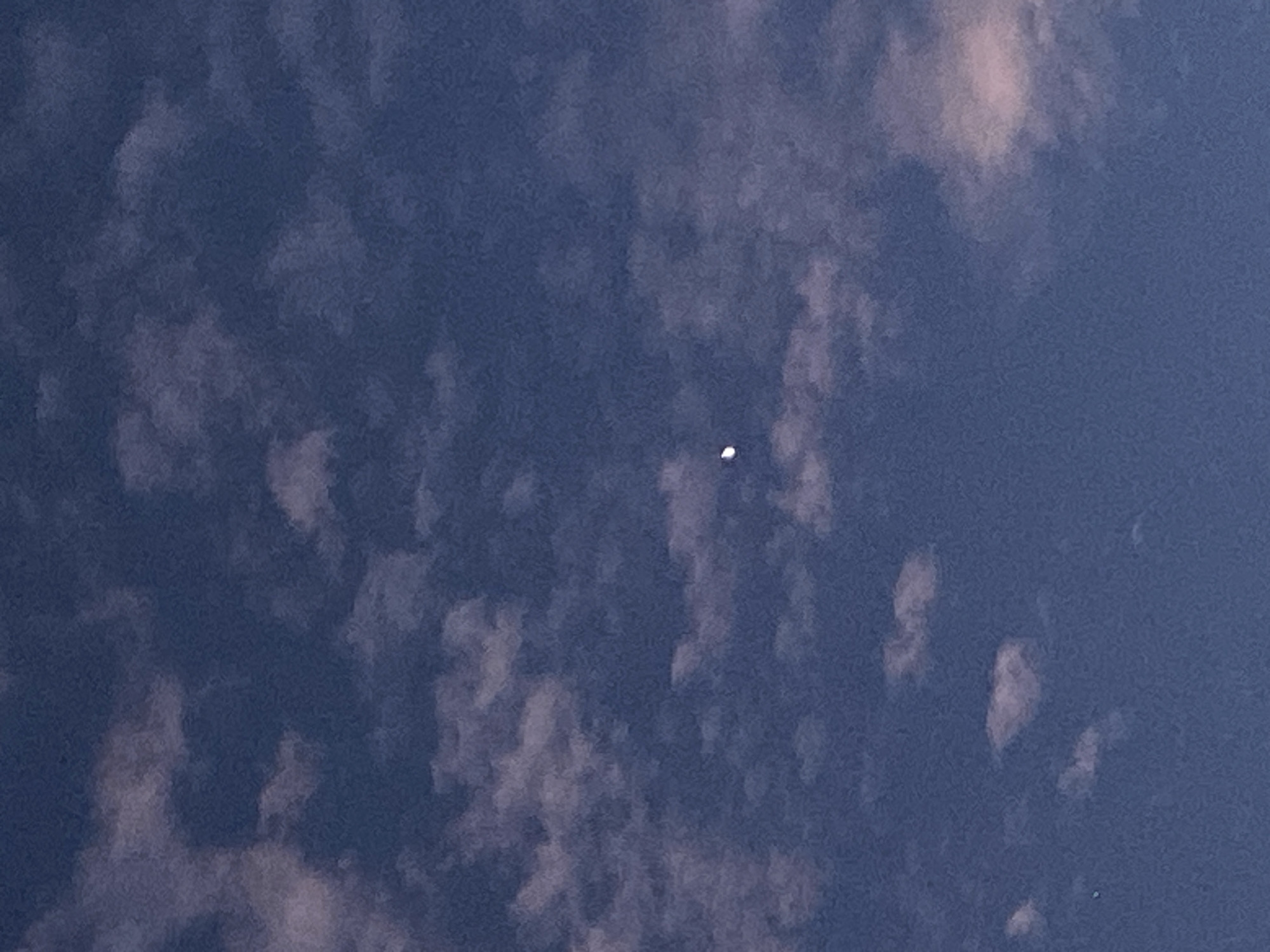 The image is a medium shot, taken from a low angle, of a deep blue sky dotted with scattered, wispy clouds. The clouds are predominantly white with hints of pink and purple, suggesting the presence of either dawn or dusk light. In the upper center of the frame, a crescent moon is visible, appearing as a thin sliver of white against the darkening sky. The overall impression is one of a peaceful, transitional moment in the day. There is no visible indication of the city of Abakaliki, Nigeria.