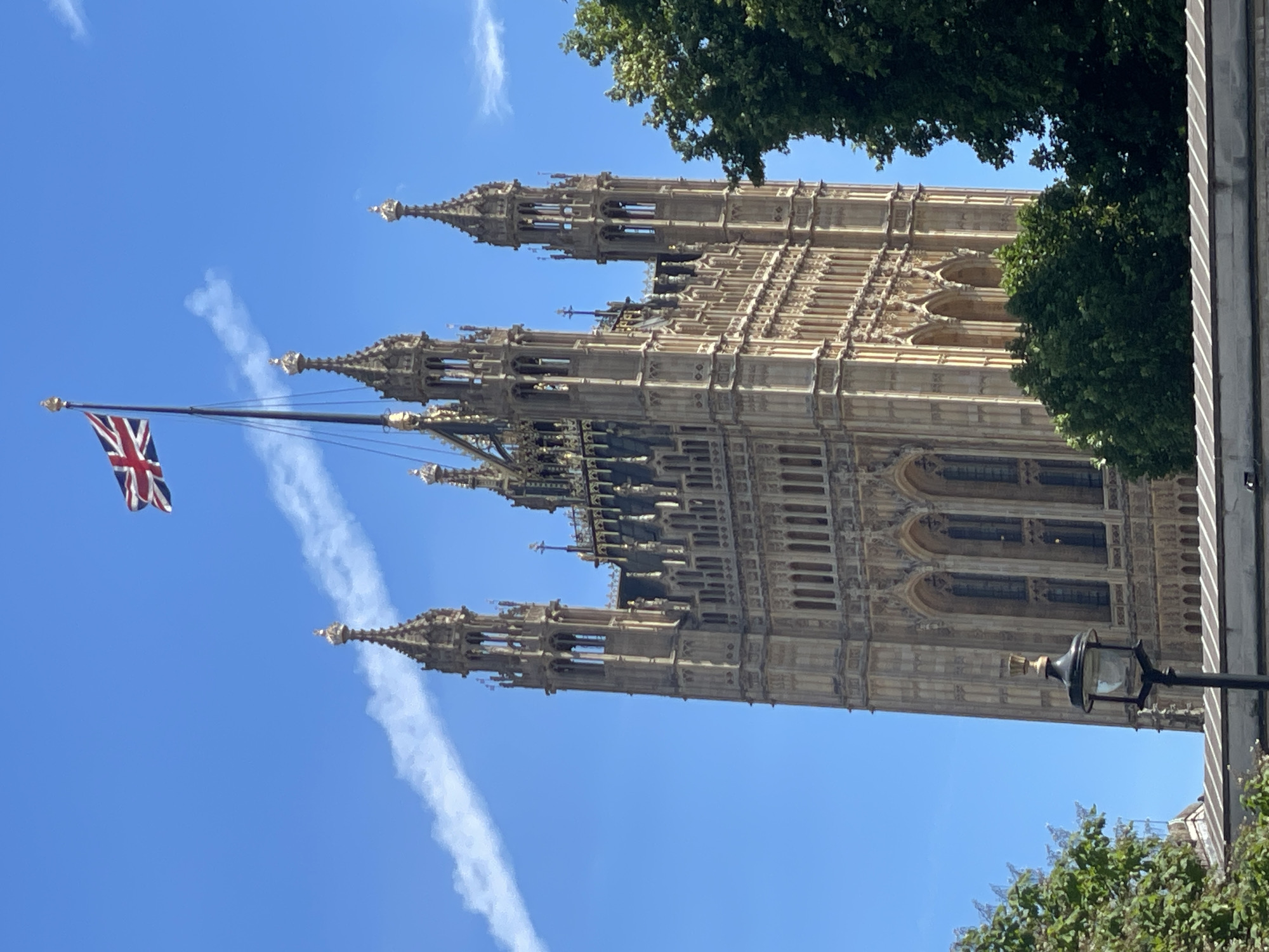 The image captures a view of the Elizabeth Tower, part of the Palace of Westminster in London, United Kingdom. The tower, constructed of limestone, is intricately detailed with Gothic Revival architecture, featuring numerous spires, pinnacles, and arched windows. A Union Jack flag is visible flying from the top of the tower. The sky is a clear, bright blue with a faint white contrail from an aircraft streaking across it. In the foreground, a dark lamppost with a globe light is positioned to the left, and lush green trees frame the right side of the tower. The lower part of the image shows the top of a stone wall or building with decorative detailing. The scene appears to be bathed in natural daylight.