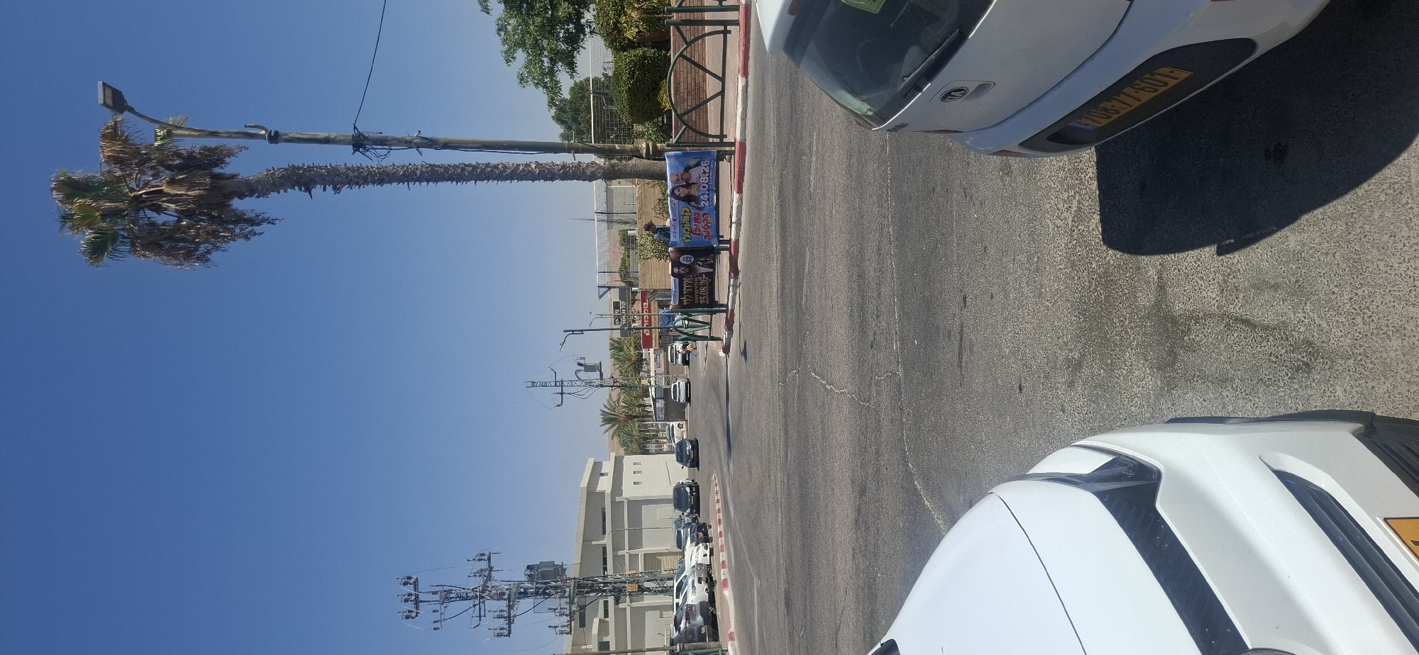 A street scene in Yerocham, Israel, on a sunny day, features a wide asphalt road with a traffic island in the foreground. Several parked cars are visible on the left side of the road, along with a large, light-colored building. Utility poles with electrical equipment are prominent. On the right side of the street, a tall palm tree stands next to a lamppost. A decorative metal fence encloses a grassy area with bushes and trees. Two banners are displayed on the traffic island, one showing two people, possibly performers or celebrities, with text in Hebrew. A person is standing near the banners. The sky is clear and blue. The perspective of the image is from a low angle, possibly from inside a vehicle, with the front of a white car visible in the lower left corner and the rear of another white car on the right.