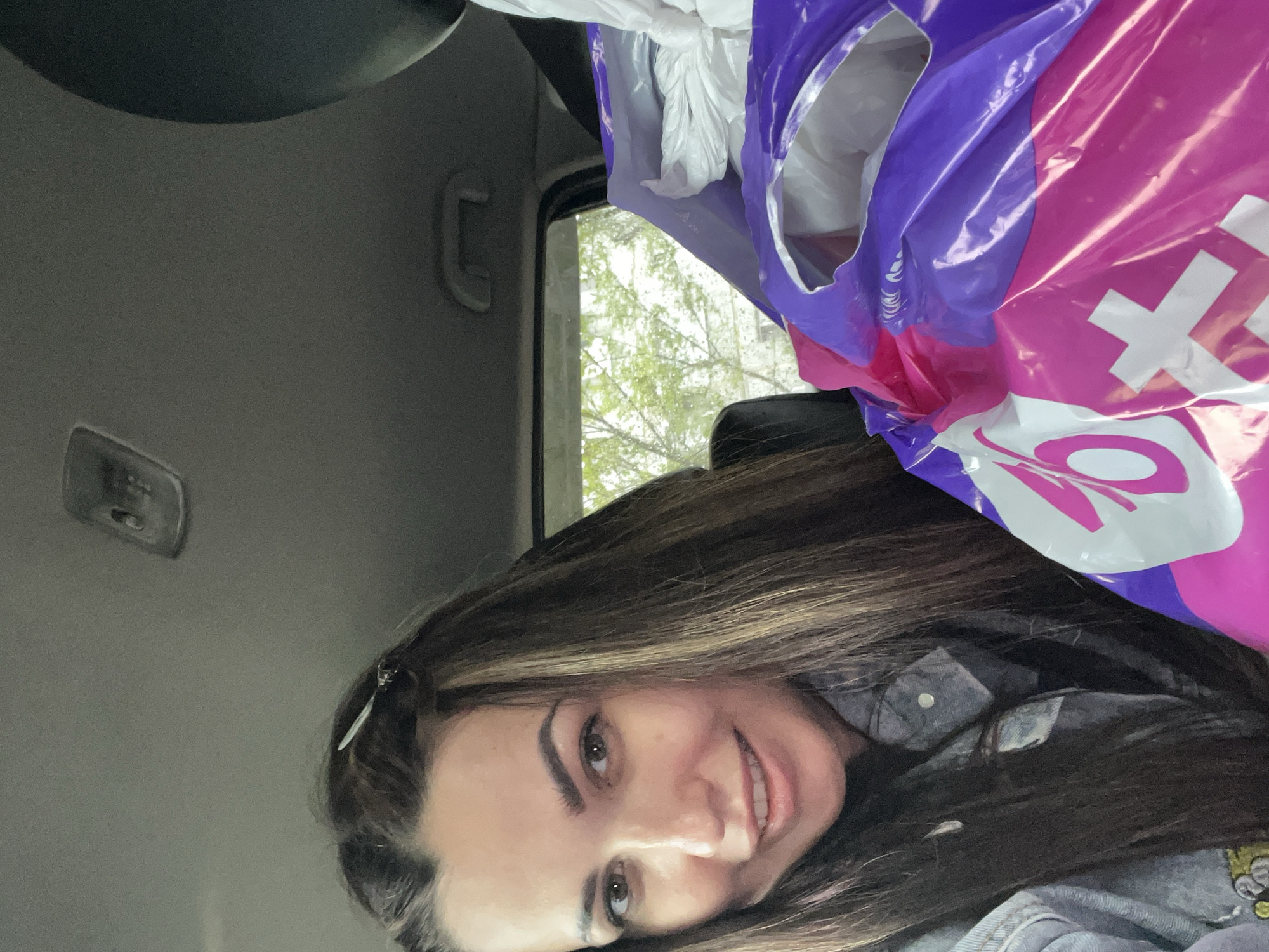 A woman with brown hair is inside a car. She is wearing a denim jacket. Above her is a purple and pink Taco Bell bag. The interior of the car is gray. Outside the car window, there are trees. The location is Unknown, Unknown.