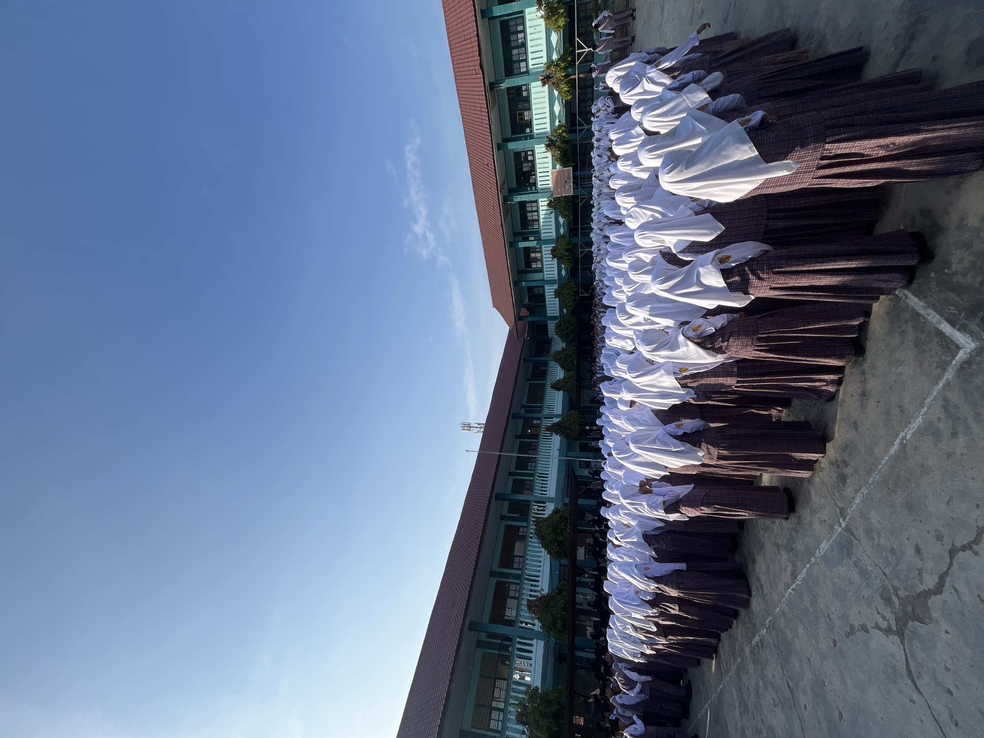 The image depicts a large group of students, likely female based on their uniforms, standing in formation on a paved schoolyard. They are dressed in school uniforms consisting of white headscarves and long plaid skirts with white tops. The students are facing away from the camera, and their postures suggest they are participating in an assembly or a similar school event. Behind the students, a two-story school building is visible. The building has a green facade with white railings and a reddish-brown roof. The sky above is clear and blue with a few wispy clouds, indicating it is daytime, likely in the morning due to the lighting. The presence of school buildings and the uniforms suggests an educational institution. Given the context of Lhokseumawe, Indonesia, this scene is likely at a school within the city. The environment is an open courtyard of a school.