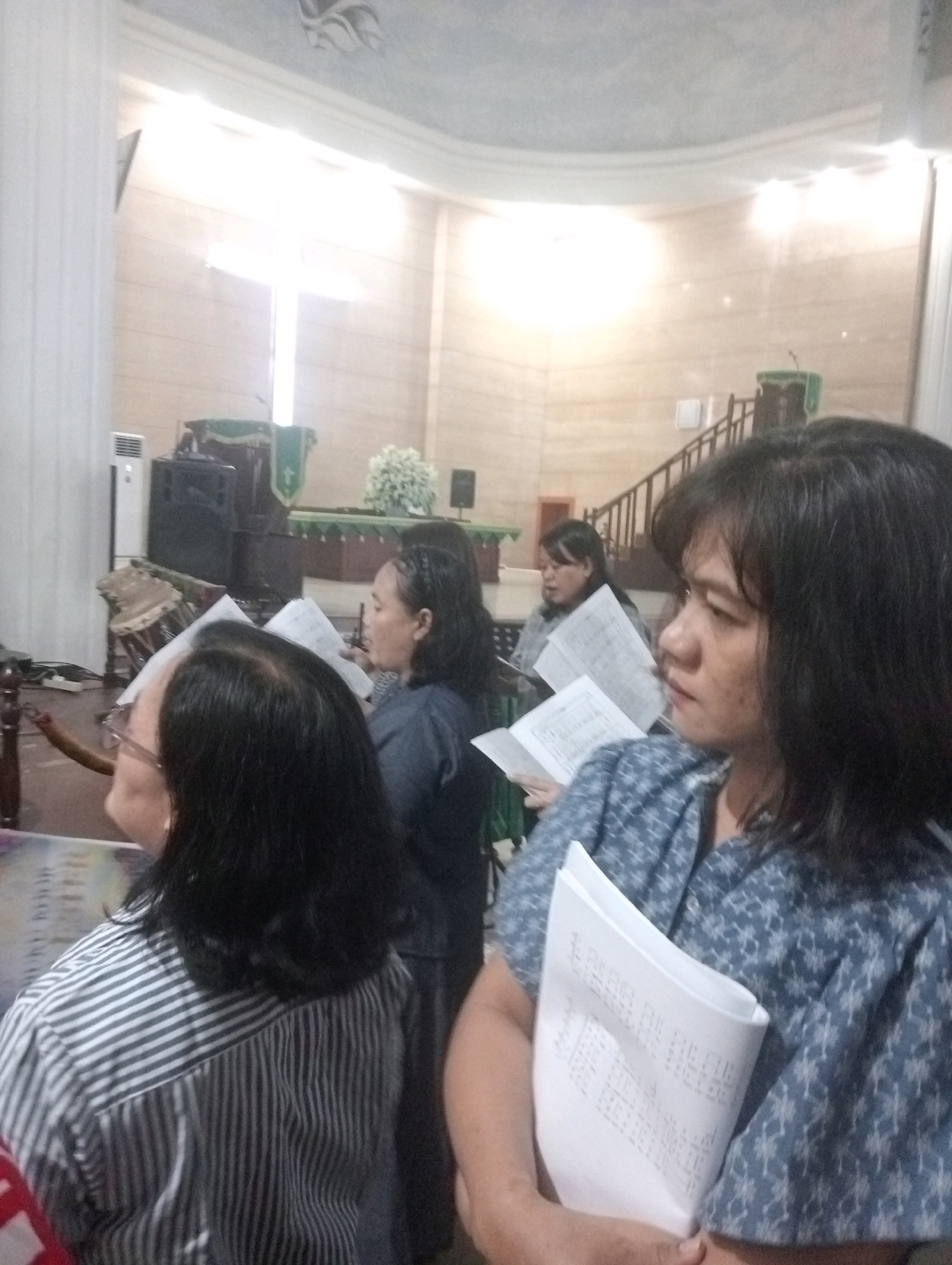 The image depicts an indoor scene, likely a church or a similar place of worship, given the presence of a pulpit with a green cloth and a cross visible in the background. Several individuals are present, some of whom appear to be holding sheet music or books, suggesting a choral or congregational activity. In the foreground, a woman with dark hair is looking to her right, holding a sheet of paper with text. To her left, another person with dark hair and glasses is also facing away from the camera. In the mid-ground and background, other individuals are visible, some also holding papers. The environment includes architectural elements like columns and a curved ceiling with decorative painting. The lighting suggests artificial illumination. The presence of sound equipment, such as speakers and a possibly a musical instrument like a drum, indicates a performance or service setting. The scene is located in Jambi, Indonesia.