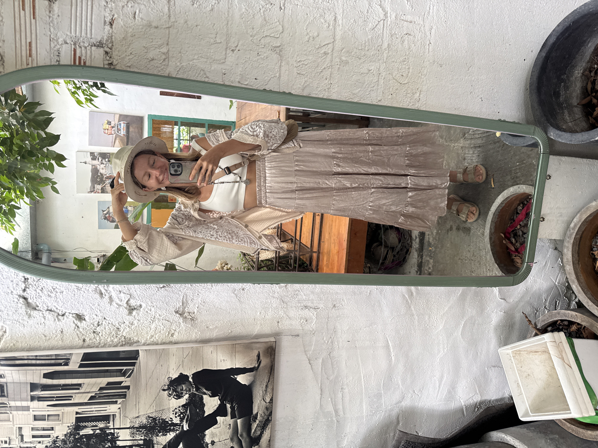 A woman is taking a selfie in a full-length mirror. She is wearing a white crop top, a beige embroidered open cardigan, a long, tiered beige skirt, a straw hat, and beige sandals. She is also wearing a crossbody bag. The mirror is leaning against a white brick wall. In the background, there is a black and white photograph of a street scene with buildings and people, and other framed pictures on the wall. There are also several potted plants and planters visible. The scene appears to be outdoors, possibly in a courtyard or garden area in Phu Thuy Ward, Vietnam.