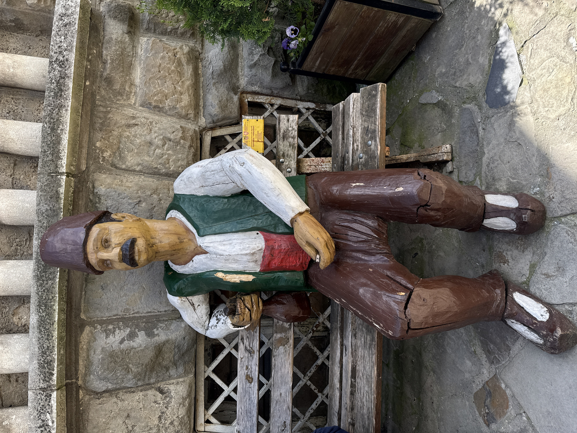 A life-sized, weathered wooden statue of a man with a mustache sits on a worn wooden bench next to a stone wall and stairs. The statue wears a brown cap, green vest with a red stripe, and white shirt, and brown pants. The bench is placed in front of a stone wall with a rusty metal grate and a small wooden planter box with a purple flower. The scene is in Veliko Tarnovo, Bulgaria.