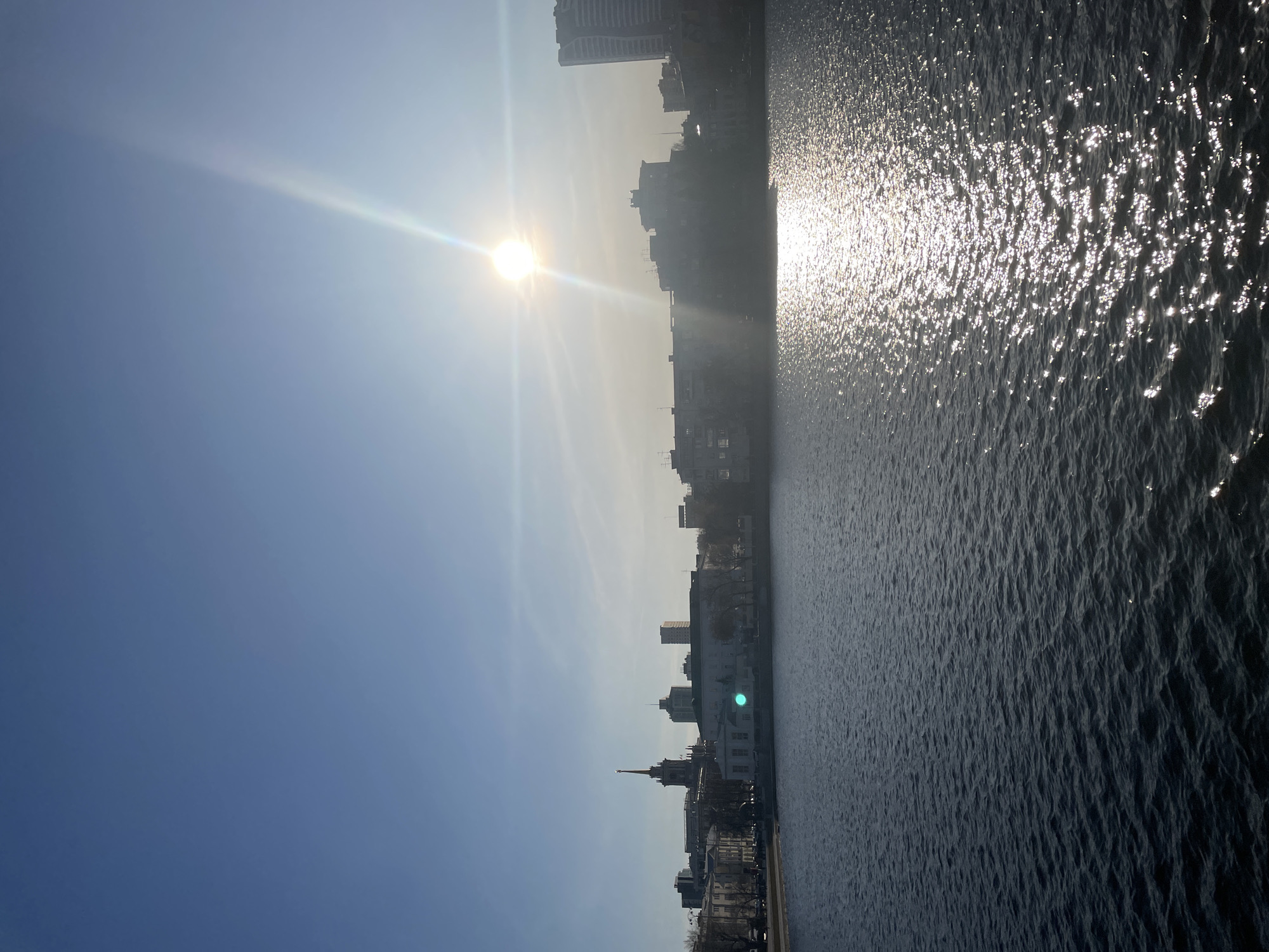 This image presents a vibrant, sunlit daytime view of a city along a body of water, likely a river or large canal, consistent with a location like Yekaterinburg, Russia. The perspective is from across the water, looking towards the city skyline.
The scene is dominated by a brilliant sun positioned in the upper center, emitting strong rays and lens flares that extend across the sky and water. The sky is a clear, bright blue, fading slightly towards the horizon due to atmospheric haze and the intense sunlight.
The body of water occupies a significant portion of the frame, its surface sparkling intensely with countless reflections of the sun. These glints create a textured, shimmering effect, particularly pronounced closer to the light source. Further from the sun, the water appears darker, with visible ripples and small waves.
Along the opposite bank, the city skyline is silhouetted against the bright sky. The buildings display a mix of architectural styles, including some older structures with distinctive spires and more contemporary, taller buildings. While details are somewhat obscured by the strong backlighting and atmospheric conditions, the overall form of the urban landscape is clearly visible.
The image conveys a clear, sunny day, with the time likely being morning or late afternoon given the low angle of the sun and the resulting glare. There is no visible text in the image.