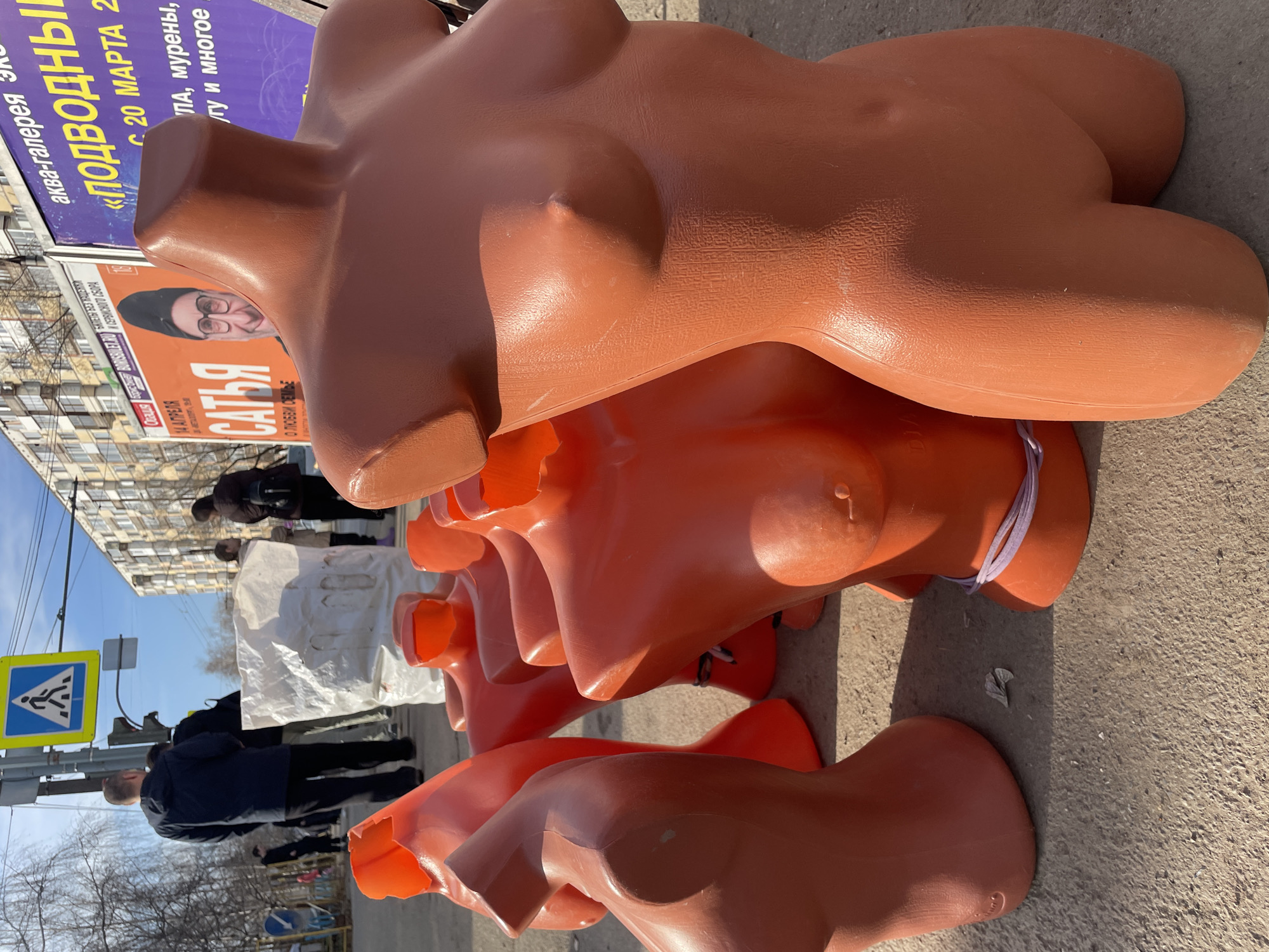 The image shows a row of orange, plastic, female torso mannequins stacked on the sidewalk in Izhevsk, Russia. The mannequins are a light orange-brown color and appear to be waiting to be set up. There is a pedestrian crossing sign in the background. People are walking on the sidewalk. The background includes buildings, some of which appear to be apartment buildings. A sign with the text "САТЬЯ" and "ПОДВОДНЫ" in Russian is partially visible.