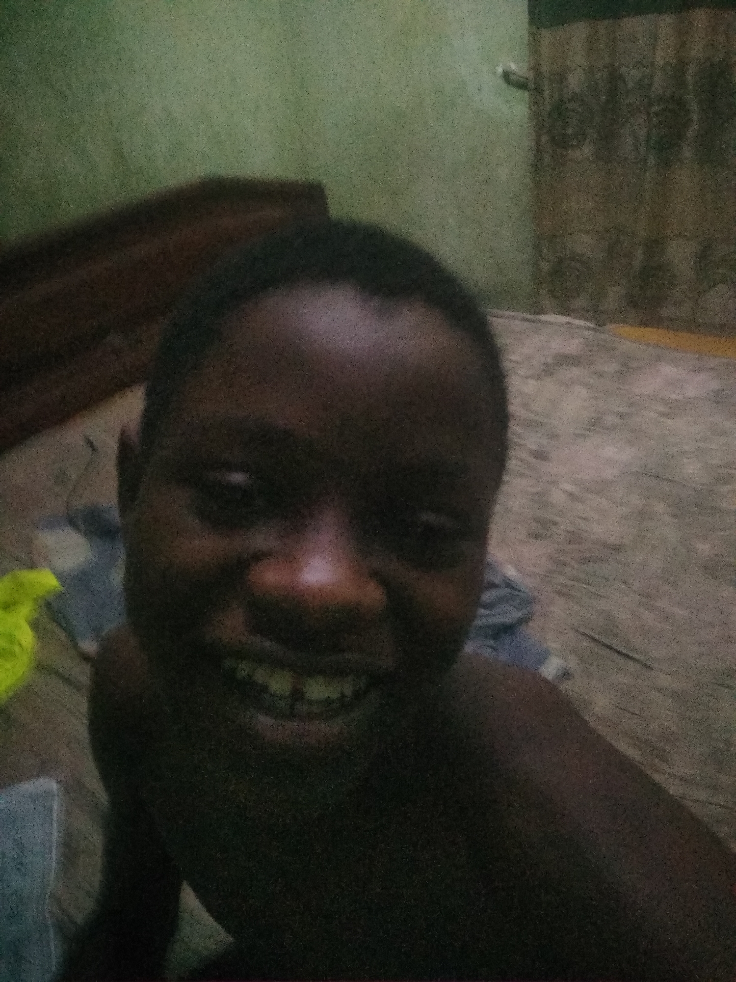 A close-up photo shows a young Black person smiling with visible teeth. The setting appears to be indoors, on top of a bed, a bed frame can be partially seen behind the subject on the left. Curtains with a patterned design are visible in the background on the right. The lighting in the room is dim. The location is not discernible from the image. The city of Shangisha, Nigeria is not discernible from the image.
