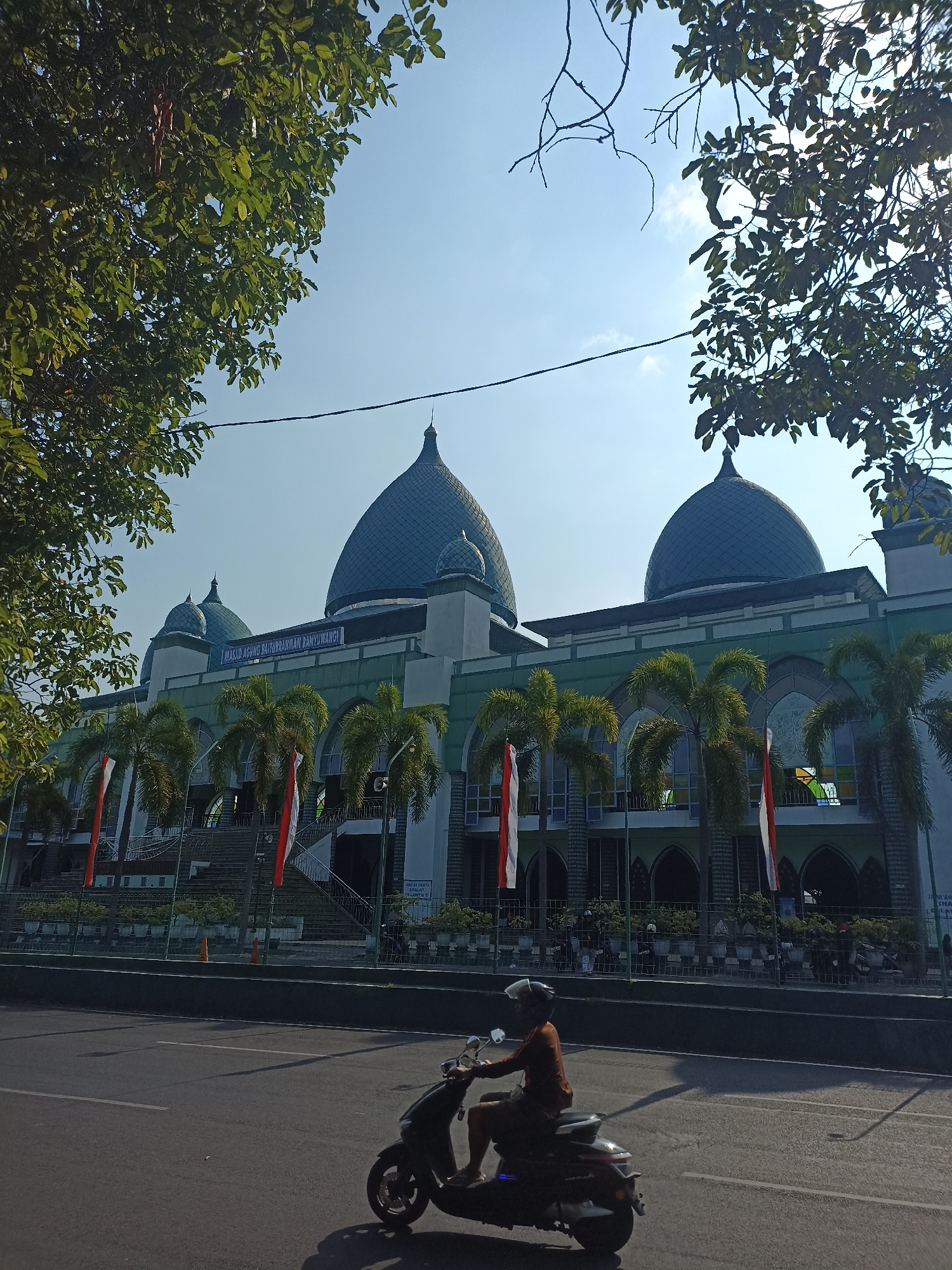 This image shows the Masjid Agung Baiturrahman, a grand mosque in Kelurahan Kepatihan, Indonesia. The mosque features a prominent large green dome and a smaller dome, with a green facade adorned with arched windows. Palm trees are planted in front of the mosque, and red and white flags are visible. In the foreground, a person is riding a dark-colored scooter on an asphalt road. The sky is clear and blue, with some sunlight casting shadows on the road and the mosque. The scene is framed by lush green trees in the upper left and right corners of the image.