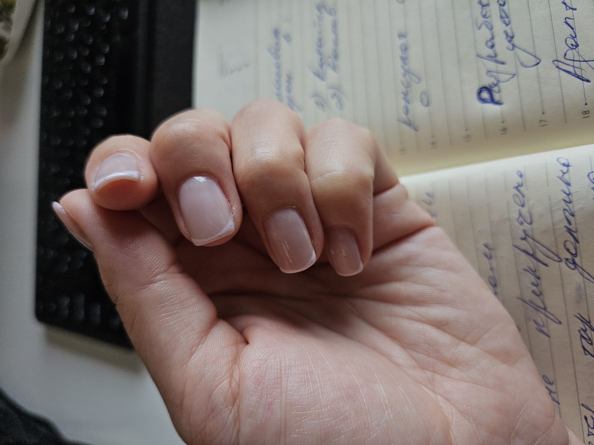 A person's left hand with a French manicure is resting on an open notebook filled with handwritten text in a blue ink. The notebook is likely indoors, possibly on a desk or table, as evidenced by the edge of a dark-colored keyboard visible in the background. The location is unknown, and there is no evidence of the city of Chaikovsky, Russia.