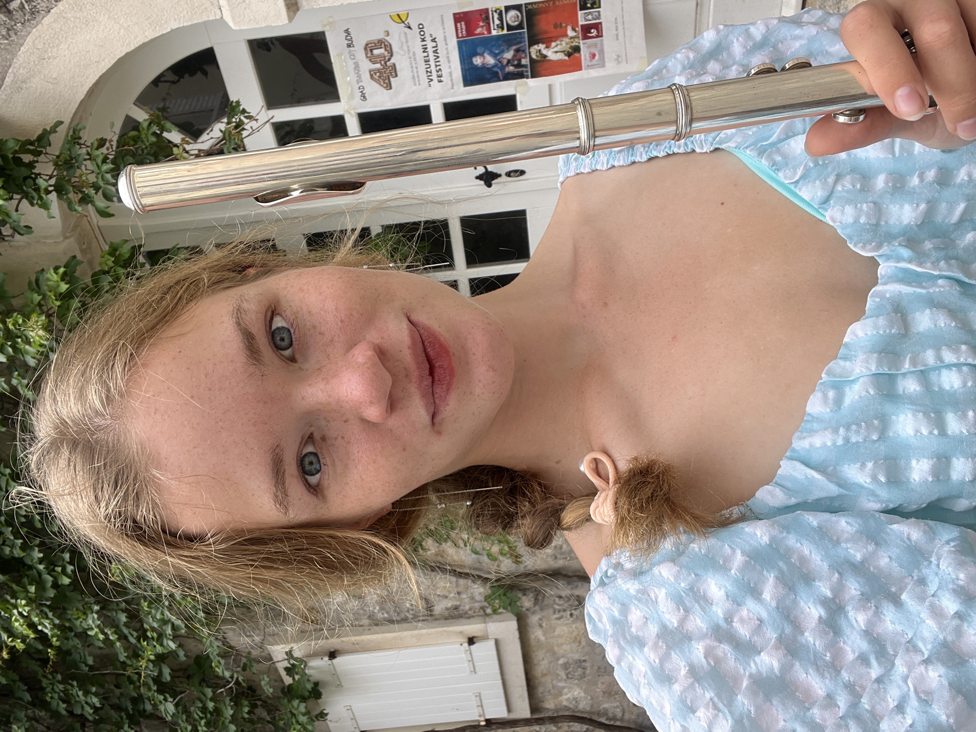 A blonde female with fair skin and freckles is holding a silver flute up to her mouth. She is wearing a light blue, textured top. Her hair is partially tied back with a peach-colored scrunchie. The background includes a white door with an arched window above it, as well as ivy growing on a stone wall. A poster is attached to the door. The poster is titled "GRAD TEATAR CITY BUDVA".