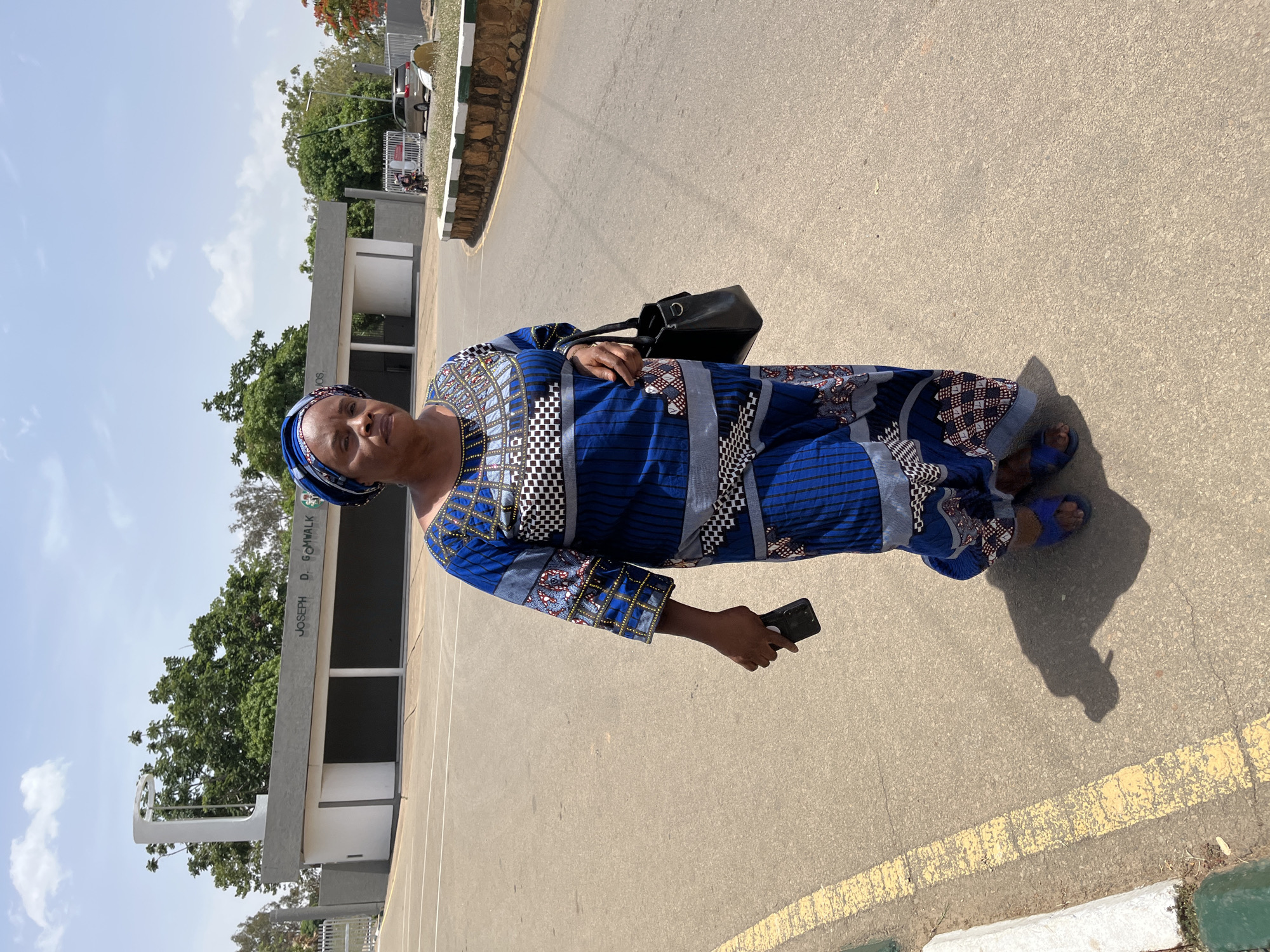 A woman stands in the city of Jos, Nigeria. She is wearing a blue, patterned, long-sleeved dress and matching head covering. She carries a black handbag on her shoulder and holds a black smartphone in her right hand. She wears blue sandals. The ground is concrete with yellow painted lines. In the background is a building marked "JOSEPH D. GOMWALK" and trees. Cars are parked in the distance. The sky is light blue. The overall setting appears to be outdoors on a sunny day.