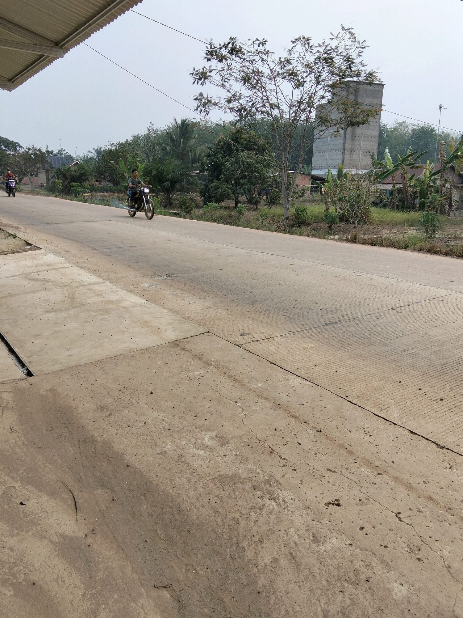 The image depicts a paved road in Perambahan, Indonesia. On the left side of the road, a section of a building's corrugated metal roof is visible. A concrete path with a drainage channel runs alongside the road in the foreground. Two individuals are riding motorcycles on the road. The rider on the closest motorcycle is a young male wearing a t-shirt, and the motorcycle is emitting a small puff of smoke from its exhaust. Another motorcycle with a rider is further down the road on the left. The background features lush greenery, including trees and banana plants, and a tall, gray, concrete building that appears to be a bird's nest or bird farm structure. The sky is overcast and hazy. The road is segmented with expansion joints.
