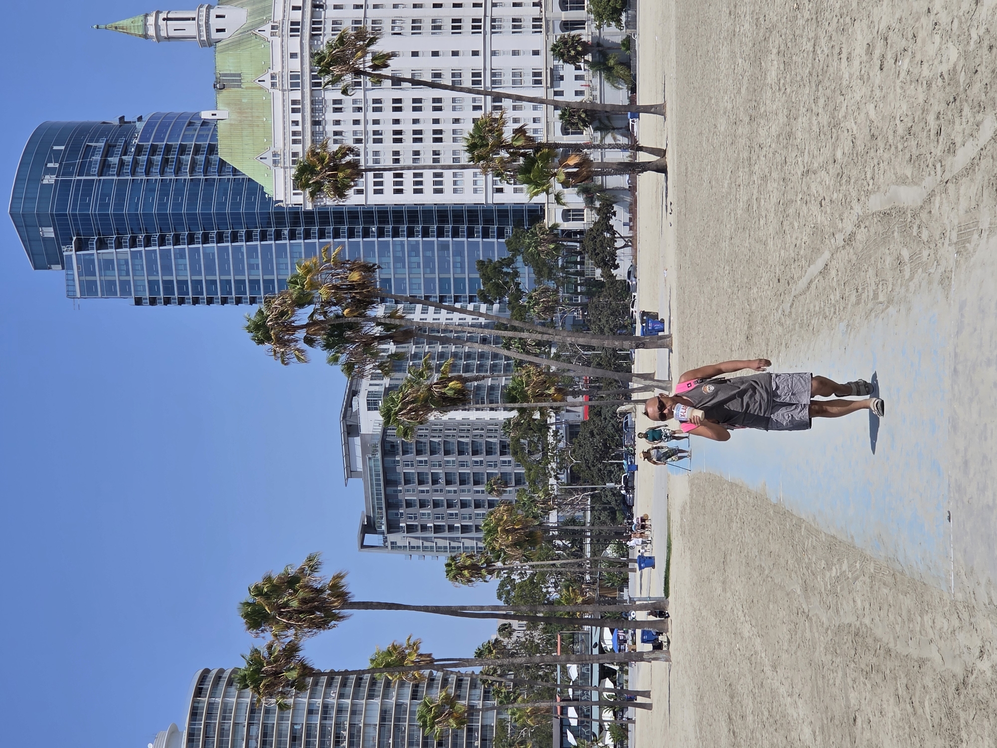 The image depicts a sunny day at the Long Beach coastline in California. In the foreground, a man walks along a blue path on the sandy beach, holding a clear cup with a straw. He wears sunglasses, a gray t-shirt, gray shorts, and sandals, with a pink backpack slung over his shoulder. Several palm trees are scattered across the beach. In the background, a cluster of modern and historic buildings forms a cityscape. Notably, a tall, dark blue glass skyscraper stands prominently next to a white building with a green, ornate roof and a spire. Other buildings and trees are visible further in the distance, and a few other people are seen walking and cycling on the beach path. The sky is clear and blue.