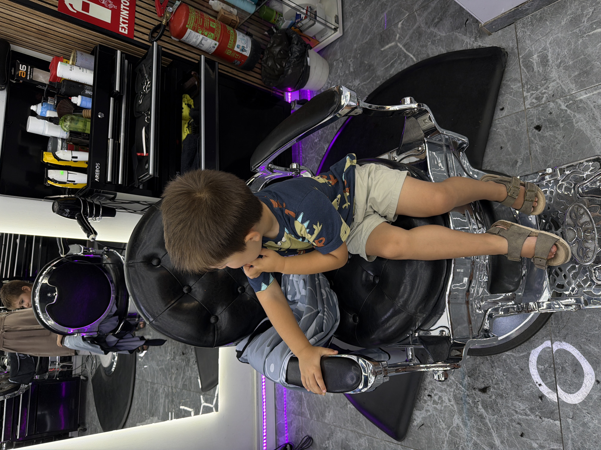 A young boy with brown hair is seated in a chrome and black leather barber chair in what appears to be a modern barbershop. He is wearing a patterned blue and orange t-shirt, light-colored shorts, and grey sandals, with a grey and blue patterned haircut cape draped over him. His head is slightly bowed, and he appears to be looking down or concentrating on something in his lap or hands, though his hands are partially obscured by the cape.
The barbershop features sleek black furniture, including a storage unit with multiple shelves holding various hair products like bottles, sprays, and brushes. Some electric hair clippers or trimmers are also visible. A red fire extinguisher is mounted on the wall above the storage unit. The floor is covered in a grey, marble-like tile, with small clippings of hair visible, indicating a haircut is in progress or has recently occurred. Purple LED strip lighting illuminates the space, particularly along the bottom edge of a mirror reflecting the room and another person (likely the barber) in the background. A hairdryer is seen on a shelf next to the boy's head.
Visible text includes "EXTINTOR" and "ESPUMA" on the fire extinguisher, and "AREBOS" on a storage drawer. Other product labels, like "ONE" and "2.6," are partially visible on bottles. The overall setting is indoors, brightly lit, with a contemporary design.