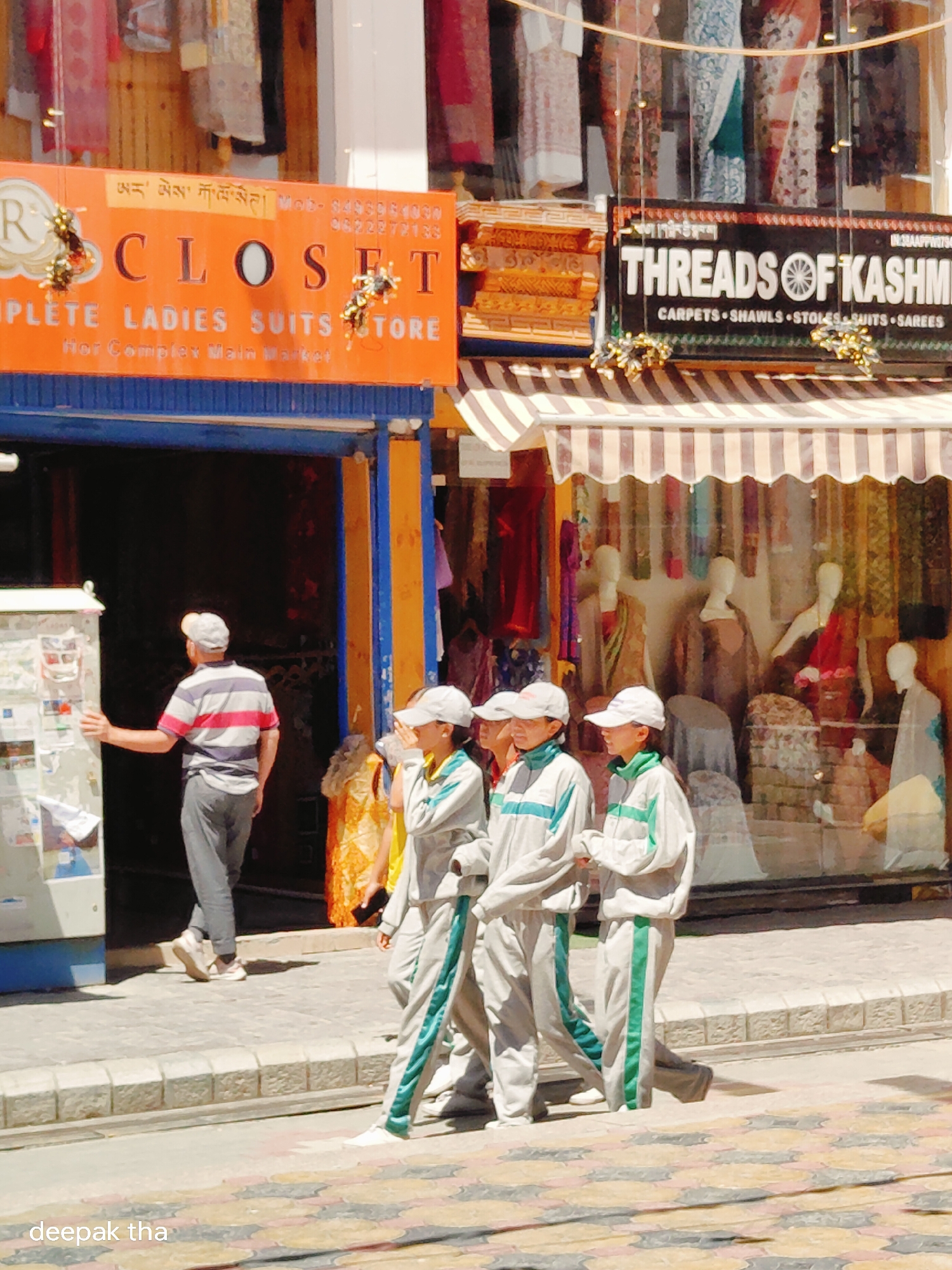 This image captures a street scene in Leh, India, likely within the main market area, as suggested by the shop signs. In the foreground, three young individuals, two females and one male, dressed in white and green tracksuits, walk from left to right. The female on the right shields her face from the sun with her hand. Behind them, a man in a striped shirt and grey pants stands near the entrance of a shop. The shop's signage, partially visible, includes "CLOSE T" and "THREADS OF KASHMIR." The environment suggests a sunny day with pedestrian traffic on a paved sidewalk. The background reveals storefronts displaying textiles and mannequins.
