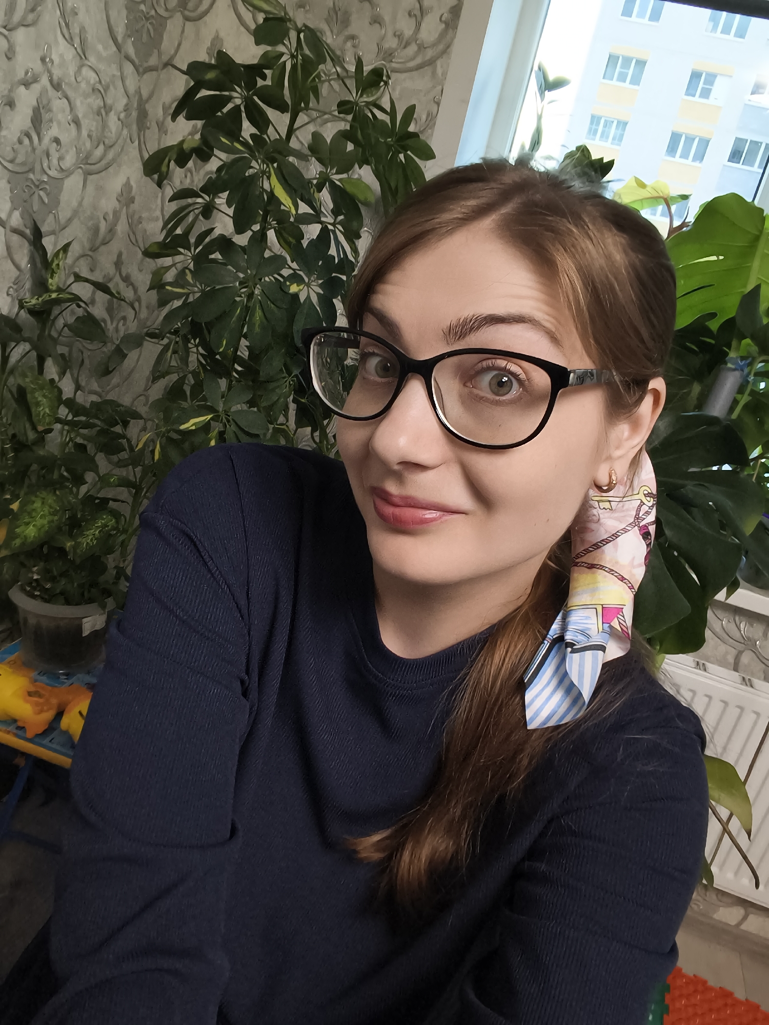 This is a medium shot selfie of a woman with light brown hair and glasses, taken indoors. She is wearing a dark blue long-sleeved shirt. Her hair is pulled back into a ponytail adorned with a patterned scarf that features stripes and a key design. She is looking directly at the camera with a slight smile and wide eyes.
Behind her, a variety of indoor plants are visible, including a large Monstera. The background also shows a wall with patterned wallpaper and a window with a view of a multi-story apartment building, suggesting an urban or suburban setting. The lighting indicates it is daytime. The woman appears relaxed and approachable, possibly in a home or office environment. There is no visible text in the image.