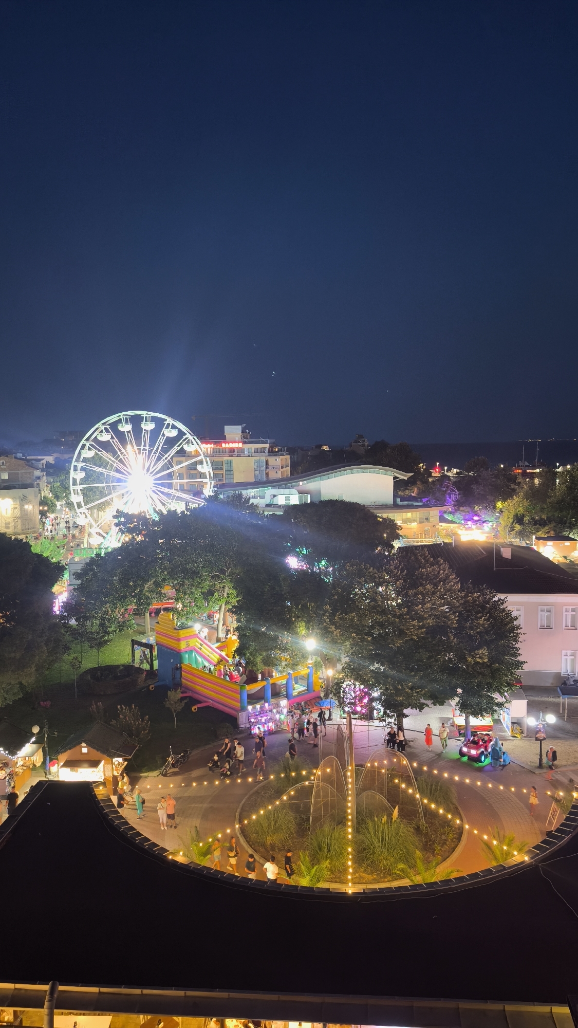 This is a nighttime aerial view of an amusement park or fairground in Pomorie, Bulgaria. In the background, a brightly lit Ferris wheel dominates the skyline, with its carriages illuminated. To the left of the Ferris wheel, a multi-story building with illuminated windows is visible, and a sign reads "PARADISE". In the middle ground, an inflatable playground, likely a bouncy castle, is brightly colored in pink, yellow, and blue. People are seen walking around the park, some gathered near the inflatable attraction and others milling about in different areas. In the foreground, a circular decorative element with an illuminated fountain and surrounding grass is lit by numerous small, warm-toned lights. Beyond this, the dark roof of a building is visible in the bottom of the frame. The overall scene depicts a lively evening at an amusement area, with a clear night sky above.
