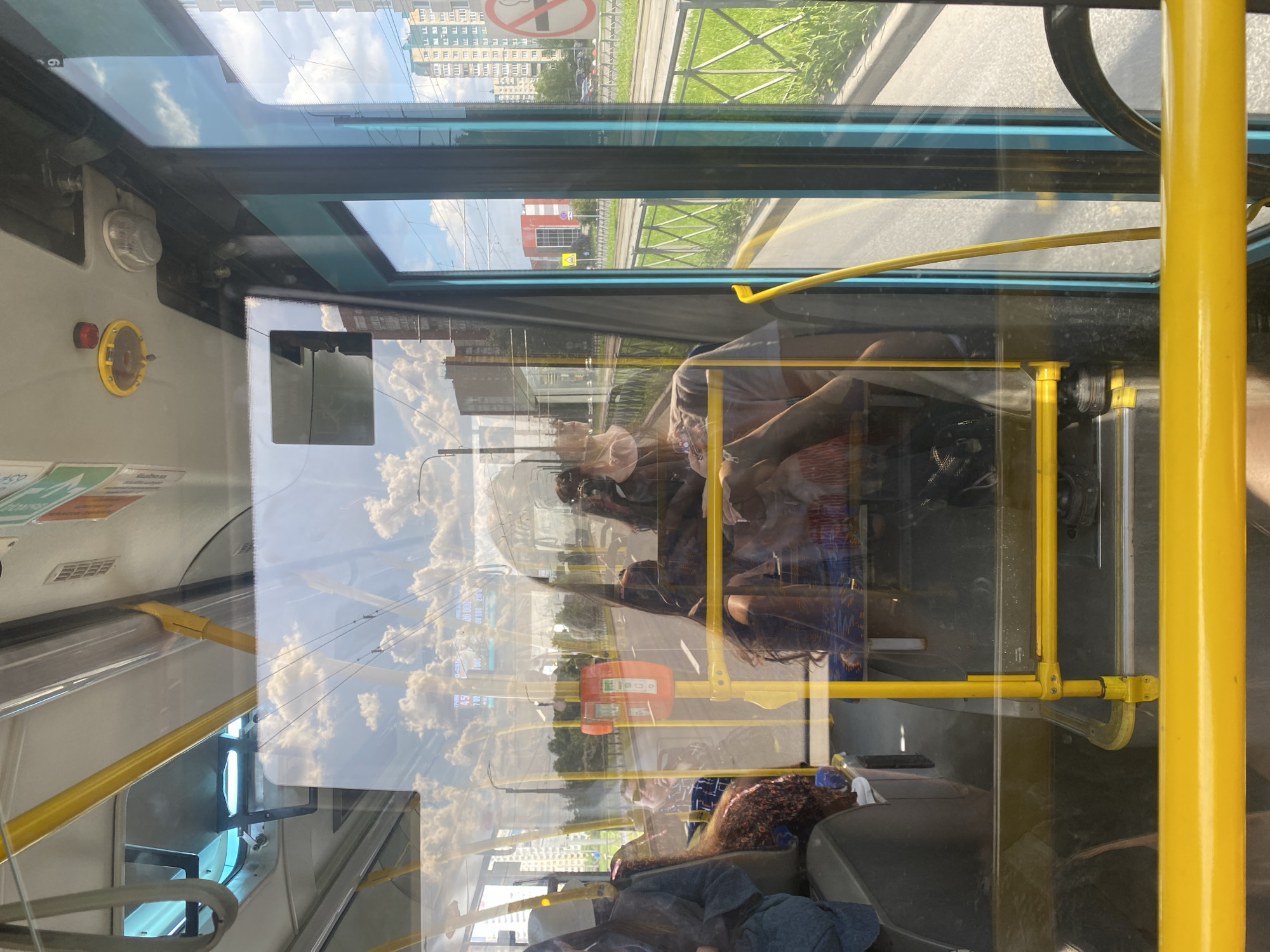 This image captures the interior of a public transport vehicle, likely a bus or tram, looking out through large windows on a bright, sunny day. The scene is characterized by strong reflections on the windows, which superimpose elements from inside the vehicle onto the urban landscape outside.
Inside, several passengers are visible, primarily seated. Their faces are partially obscured by reflections and angles, but some appear to be wearing face masks. The interior features prominent yellow handrails and poles, providing support for passengers. A yellow emergency stop button with a red center and an orange payment terminal/validator are affixed to the poles. A digital screen displays numbers such as "100," "51," "45," "700," and "0000.00," possibly indicating route information, time, or fare. Other partial texts like "ECO" are visible on interior stickers.
Through the windows and integrated with the reflections, an urban environment is seen. This includes tall, multi-story residential buildings, green grassy areas, a road, and tram tracks. Overhead, power lines stretch across a clear blue sky dotted with white clouds. The bright daylight is a notable detail, highlighting the reflections and the outdoor scenery.
A "no smoking" sign, depicted as a red circle with a crossed-out cigarette, is clearly visible, aligning with the location context of Saint Petersburg, Russia. The image conveys the experience of riding public transport through a city, with the interplay of interior and exterior views creating a dynamic perspective.