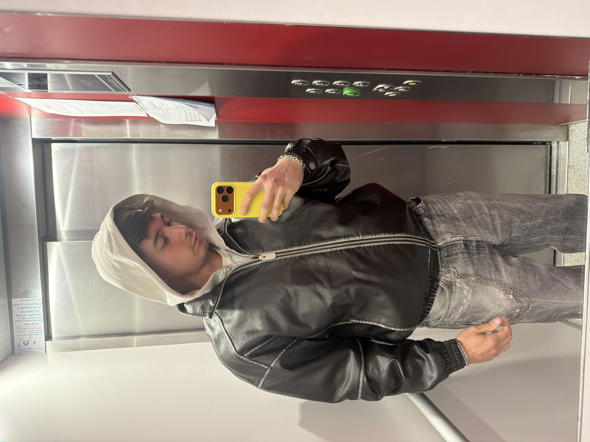 A young man is captured in a full-body mirror selfie, likely inside an elevator in Sofia, Bulgaria. He is dressed in a black leather jacket featuring prominent white contrast stitching, worn over a light-colored hooded sweatshirt. His bottom wear consists of light grey or faded black jeans, adorned with a silver chain accessory that dangles from the waist. A matching silver chain bracelet is visible on his left wrist. He holds a bright yellow smartphone, equipped with a triple-lens camera, to take the photograph. His dark hair is visible beneath the hood, and his expression is neutral as he looks down at the phone.
The setting is characterized by the reflective stainless steel walls of the elevator, a horizontal red panel, and a control panel featuring numerous buttons, one of which is illuminated with a green light. Several crumpled receipts are tucked into a narrow gap above the control panel. On the left wall of the elevator, a white informational sheet is affixed, displaying a mix of Cyrillic and Latin text, some of which is mirrored. Identifiable fragments from the text include "ГОД И БЛЕНЕН ВТ" and numbers such as "39 32 1609". The image conveys a casual, everyday moment of self-documentation within an indoor public space.