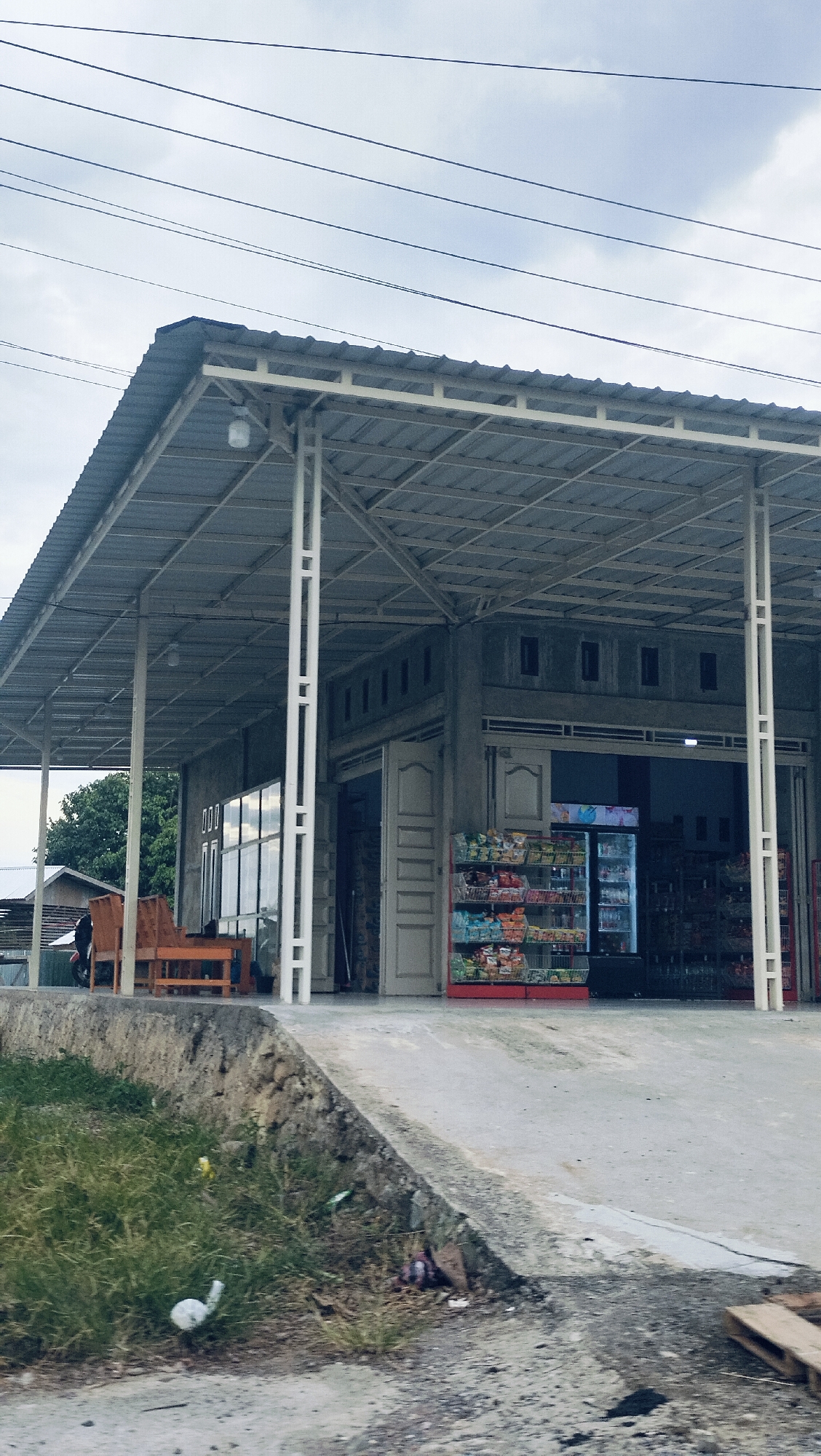 The image captures a modest, open-front store under an overcast sky in what appears to be a rural or semi-urban setting, consistent with the Lonbaroh, Indonesia context. The main subject is a single-story concrete building with a wide, corrugated metal roof that extends forward, creating a covered porch or waiting area supported by white metal columns.
Inside the store's open entrance, racks filled with various packaged snacks are visible, along with a glass-front refrigerator stocked with drinks. On the left side of the covered area, a set of light-brown wooden benches and a table provide seating. A single person, dressed in dark clothing, is partially visible sitting on one of these benches. A dark motorcycle is parked nearby.
In the foreground, a concrete slope leads up to the store. To the left of this slope, a patch of green grass is visible, containing some light-colored litter. On the right, the ground is concrete and dirt, with a wooden pallet lying near the bottom right corner. Several black power lines stretch across the cloudy sky in the upper portion of the frame. The overall impression is a functional, roadside convenience shop. No discernible text is clearly visible on any signs or products.