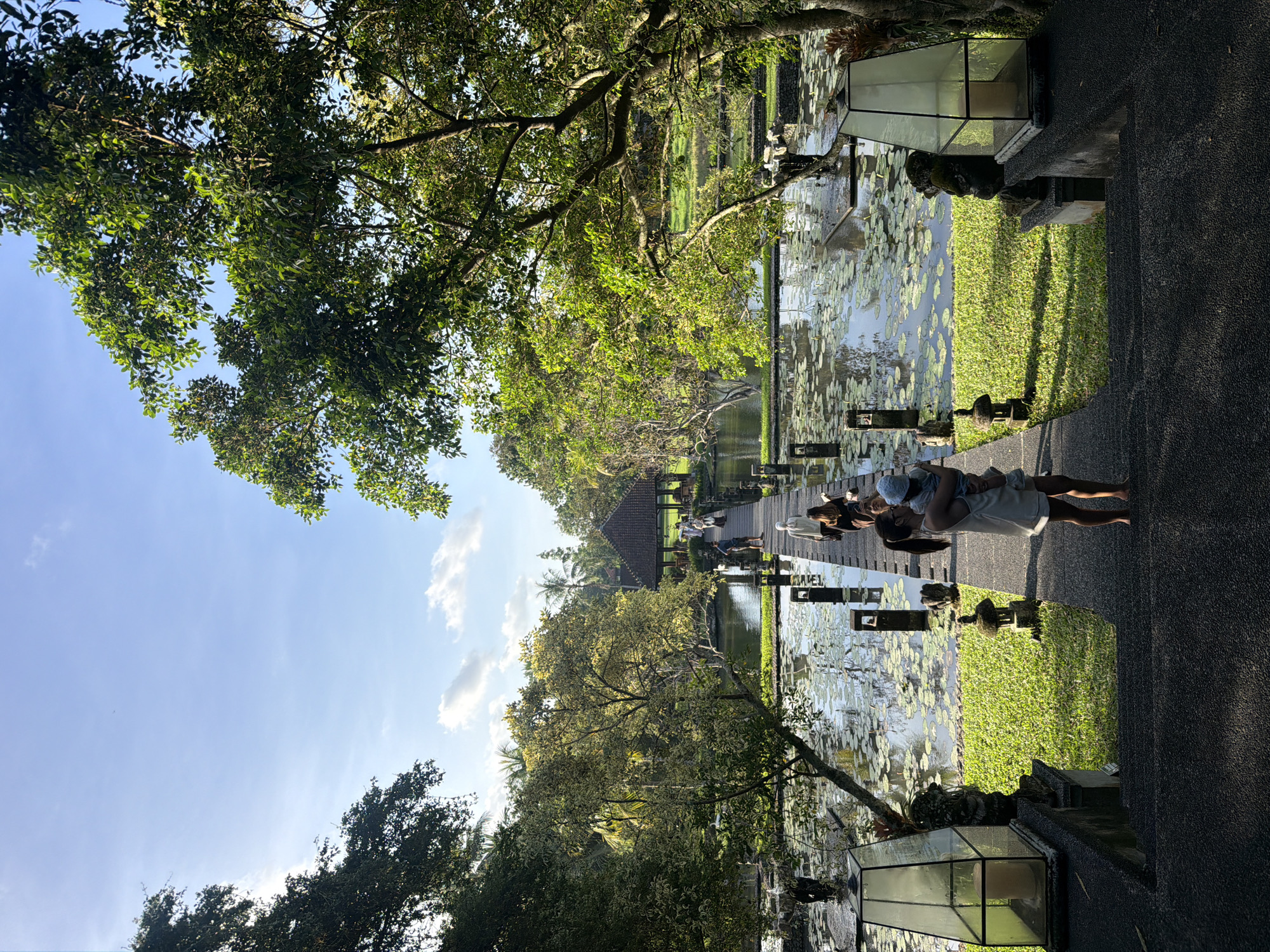 The image shows a view of Saraswati Temple in Sukawati, Indonesia, taken from an elevated position. In the foreground, part of a stone walkway is visible, with a woman holding a child standing on it. There is a pond covered with lily pads on the left side of the walkway. A stone structure and well-manicured green grass are along the side of the pond. A wooden bridge extends across the pond, leading towards a traditional Indonesian-style pavilion. Other people are on the bridge and in the pavilion. The sky is visible through the trees, indicating a sunny day. A lantern is visible on the left, and some ornamental stones along the walkway.