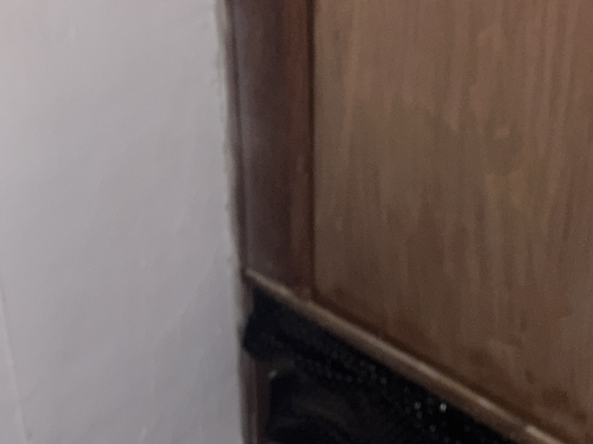 The image shows a close-up, slightly blurred view of a wooden surface, likely a door or cabinet, with a black object positioned along its top edge. The wooden surface has a visible grain pattern and appears to be a medium-brown color. Above the wood, a portion of a black, textured object is visible, which could be a bag or a piece of fabric. To the right of the wooden surface is a plain, light-colored wall, possibly painted white or a light gray. The lighting is dim, casting soft shadows. The scene is an interior shot. The location in Santa Ana, Venezuela is not determinable from the image content.
