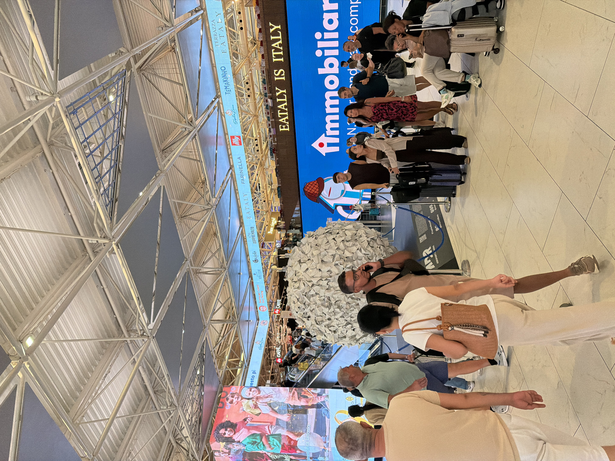 The image depicts a busy interior scene, likely at Fiumicino Airport in Italy, evidenced by the "Aeroporti di Roma" (ADR) logo visible on a blue barrier in the background. The environment is characterized by a large, open space with a high, industrial-style ceiling featuring exposed metal beams and grey panels. A prominent, large LED screen displays the text "EATALY IS ITALY" in yellow letters on a vibrant blue background, accompanied by a graphic of a house with a red hat. Above this, a row of smaller signs advertises various brands, including "EATALY," "FARINELLA," "Illy," and "TEMAKINHO."
In the foreground, several individuals are visible, most of whom appear to be travelers. On the left, a man in a tan t-shirt and white shorts is seen from the back. To his right, a woman in a white t-shirt and beige pants, carrying a woven handbag, walks away from the camera. Next to her, a man in a light green polo shirt and dark shorts looks to his left. Further back, a man with glasses and a tan shirt is on his phone, standing near a large, spherical sculpture made of what appears to be crumpled white paper.
A group of people are gathered near the LED screen, some with luggage. Several individuals are looking at the screen or engaging in conversation. Escalators are visible in the background, indicating a multi-level area. The floor is tiled with large, light-colored square tiles. The overall impression is of a modern, commercial space within a transportation hub, with an emphasis on Italian food and culture as suggested by the Eataly branding.