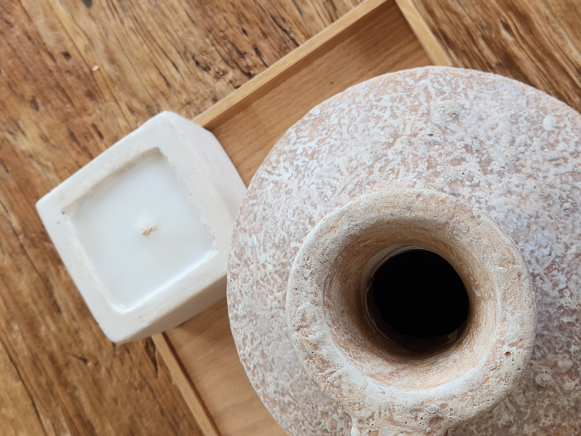 The image shows a close-up, overhead view of two decorative items on a wooden surface. In the foreground, the neck and upper portion of a large, textured, light-colored amphora are visible. The amphora has a rough, porous surface with areas of white and pinkish-brown discoloration. Its opening is a dark, circular void. To the right and slightly behind the amphora, a square, white candle sits on a light-colored wooden tray. The candle is unlit and appears to be made of wax. The background consists of a distressed wooden surface with visible grain and knots. The overall scene is stationary and staged, likely for decorative purposes. There are no people or animals present, and no actions or interactions are depicted. The location in Maitland, United States of America is not discernible from the visual information provided.