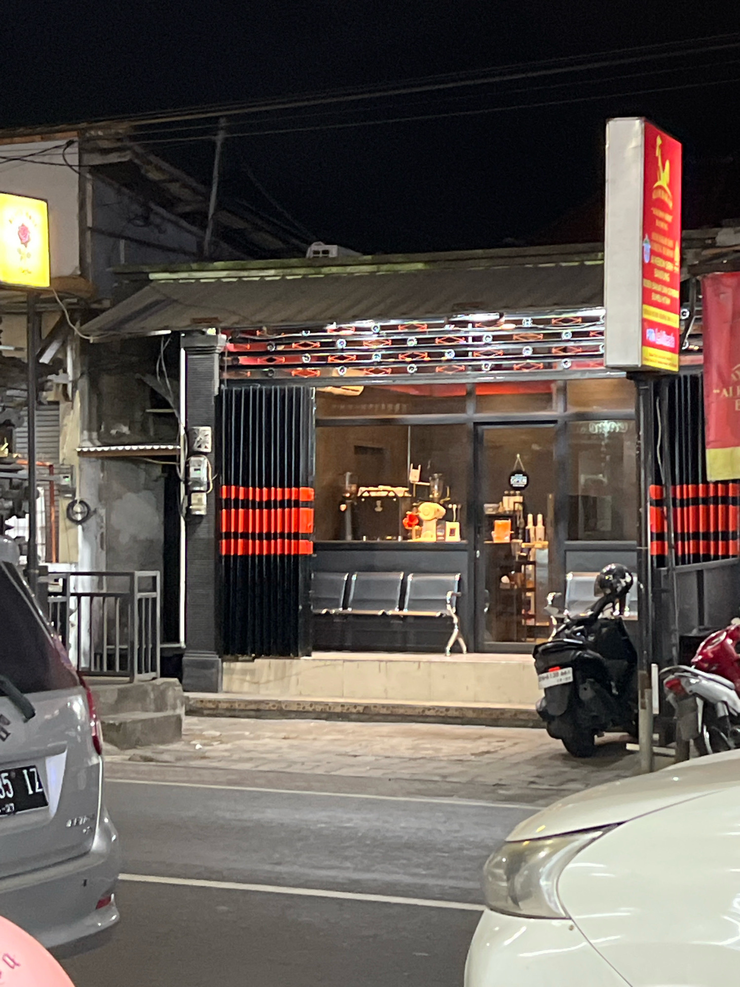 The image captures a street scene at night in Denpasar, Indonesia. The focal point is the facade of a commercial establishment, likely a cafe or restaurant, with large glass windows revealing the interior. The building's entrance is flanked by a black and orange striped gate, closed for the night. Inside, rows of empty seats are visible, suggesting it was previously occupied or is prepared for customers.
Above the entrance, a series of lights illuminates the area. A vertical red and white sign with yellow lettering stands prominently to the right of the entrance, likely advertising the business. To the left, a square yellow sign with a pink rose motif is also illuminated.
The street in front of the establishment is paved and appears to be a public road. A silver car is parked on the left side of the frame, its license plate partially visible. A black motorcycle is parked in front of the cafe, and a red motorcycle is partially visible to its right. The overall environment is urban, with overhead wires and other signs of city infrastructure present. The lighting suggests it is nighttime, creating a somewhat subdued atmosphere.