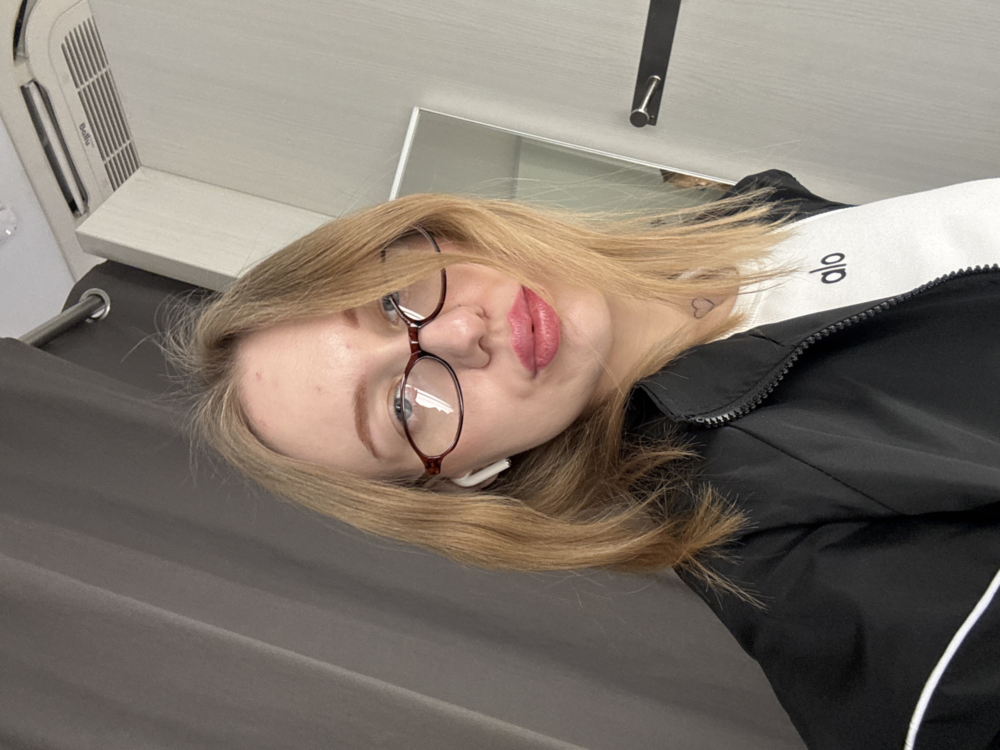 An image taken in Rostov-on-Don, Russia, depicts a female individual, appearing to be in her twenties or thirties, captured in a selfie. She has shoulder-length blonde hair, wears dark-framed, cat-eye style eyeglasses, and a white wireless earbud in her left ear. Her lips are coated with pink/red lipstick and slightly pursed. A small heart outline tattoo is visible on the left side of her neck. She is dressed in a black zippered jacket, displaying a white stripe on the left arm, over a white ribbed top bearing the black lowercase text "alo." The background on the left consists of dark grey draped curtains with a visible silver curtain rod. The right side of the background features light grey wall paneling, with an inverted "Ballu" brand air conditioning unit mounted at the top right, a vertical section of a mirror in the mid-right, and a dark horizontal bar with a silver handle fixed to the wall on the lower right. The individual is looking directly at the camera.