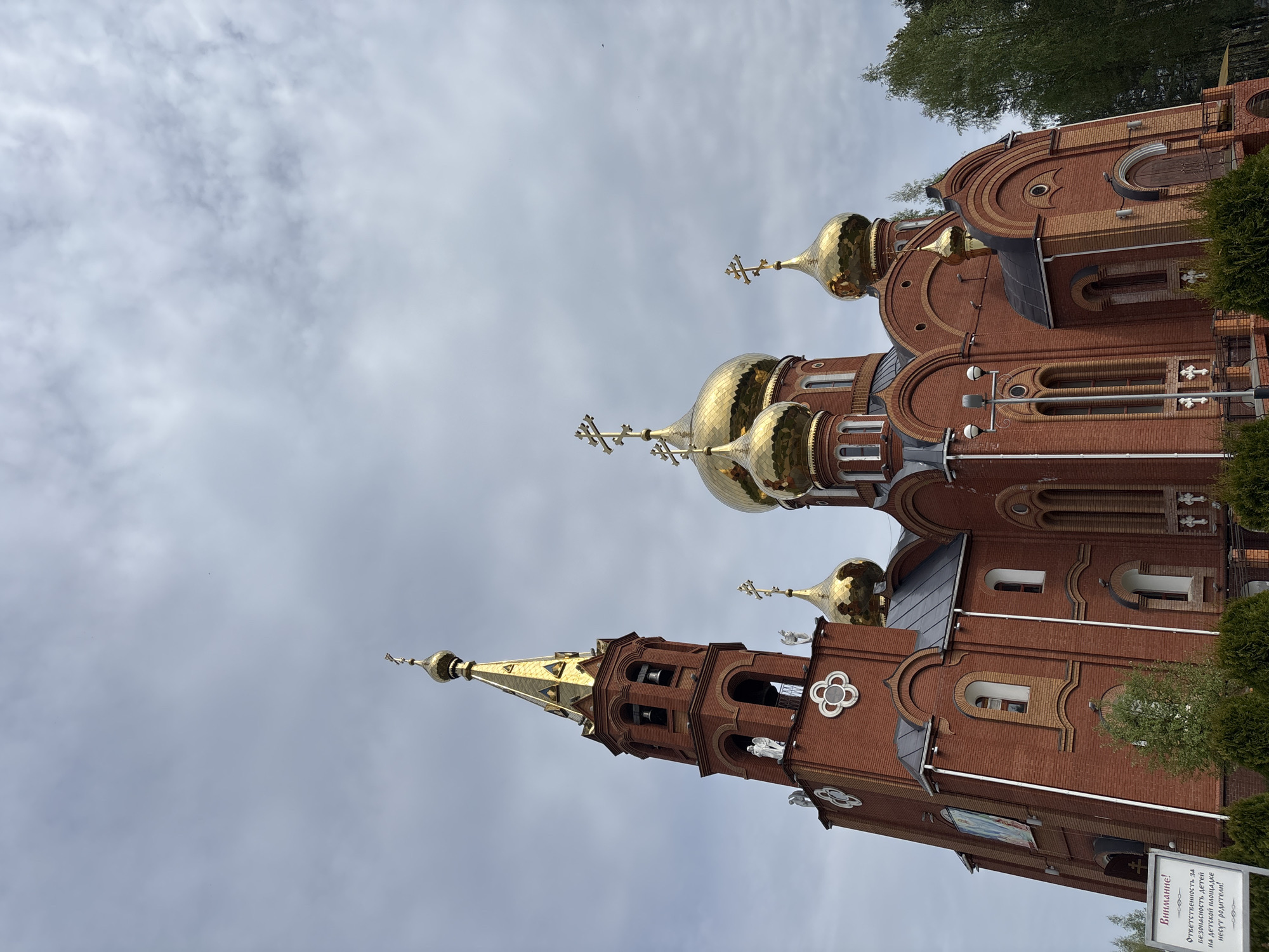 The image shows a brick church in Chaikovsky, Russia. The church features multiple golden domes and a prominent bell tower with a golden spire. The sky is overcast. A sign in Russian is visible in the foreground. The church architecture includes arched windows and decorative elements. Trees are visible in the background. The perspective is from a low angle, looking up at the church.