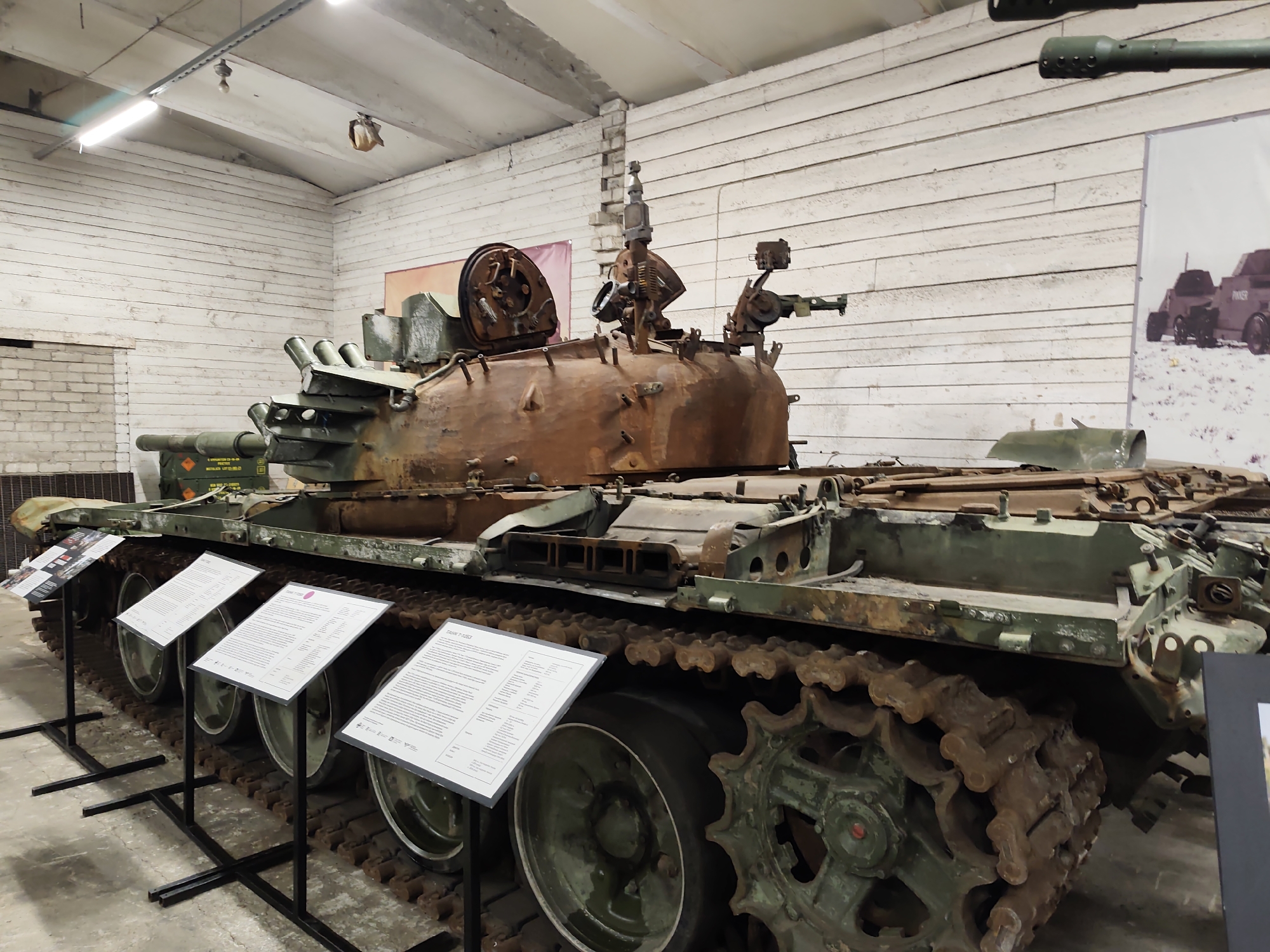 The image shows a rusty, damaged tank, likely a Soviet-era T-54 or T-55, displayed indoors. The tank is positioned on its tracks, with its turret and gun barrel visible. The interior of the building appears to be a museum or exhibition space, with white wooden paneling on the walls and fluorescent lighting. Several informational displays are placed in front of the tank. The tank itself shows signs of wear and tear, with significant corrosion and missing components. In the background, a poster featuring other military vehicles is mounted on the wall. The tank is the primary subject of the image, and the setting is a museum environment. The location is the Viimsi Open Air Museum in Viimsi, Estonia, which is known for its collection of Soviet military equipment.