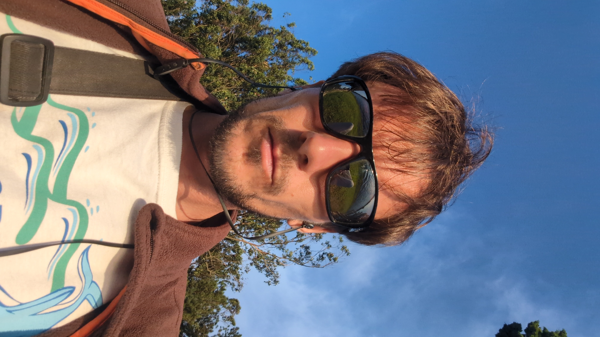 A close-up, low-angle selfie shows a man with brown, windswept hair and a light beard, wearing black sunglasses and a brown zip-up jacket over a white t-shirt with a graphic print of blue and green waves. He has black earbuds in, with the wires hanging down. The sunglasses reflect a grassy area and what appears to be a body of water. Behind him, the sky is a clear, bright blue with a few wisps of clouds. Green foliage, likely trees or bushes, is visible on either side of the frame, with the top of a tall evergreen tree visible in the upper right corner. The scene appears to be outdoors, possibly in a park or scenic area with natural surroundings. There is no specific indication that this scene is located in Da Lat, Vietnam.