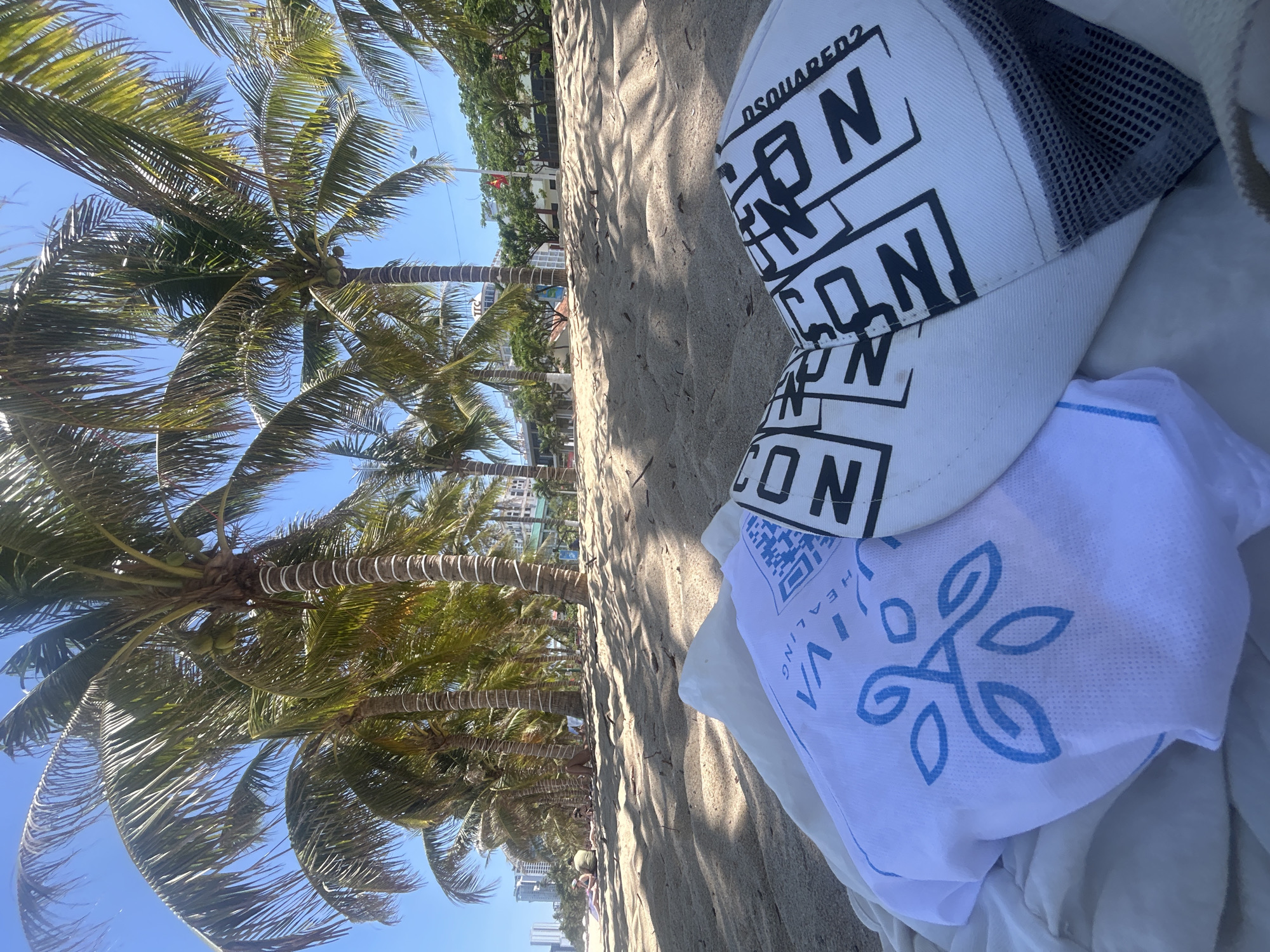 The image shows a sandy beach in Nha Trang Ward, Vietnam, lined with palm trees. A white Dsquared2 baseball cap with the word "ICON" printed multiple times and a white towel with a blue emblem are placed on the sand. People are scattered along the beach. The sky is clear and blue.