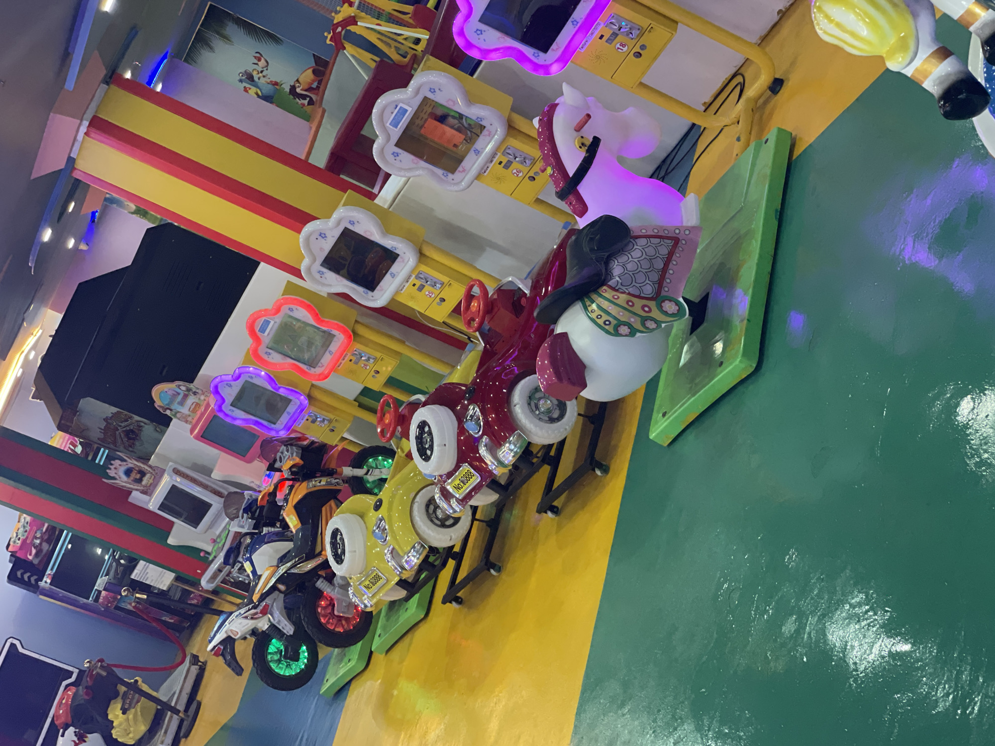 The image shows an indoor arcade in Phu Thuy Ward, Vietnam. Several children's amusement rides are lined up, including two motorcycle-shaped rides with colorful wheels, a yellow car-shaped ride, and a red car-shaped ride. In the foreground, a white and pink unicorn ride is visible, illuminated by purple LED lights. To the right, another amusement ride, possibly a horse, with yellow and white accents is partially visible. In the background, arcade machines with screens and colorful lights are present, along with decorative pillars painted in red, yellow, and green stripes. The floor is a combination of green and yellow surfaces. The overall scene depicts a vibrant and playful environment typical of an arcade.