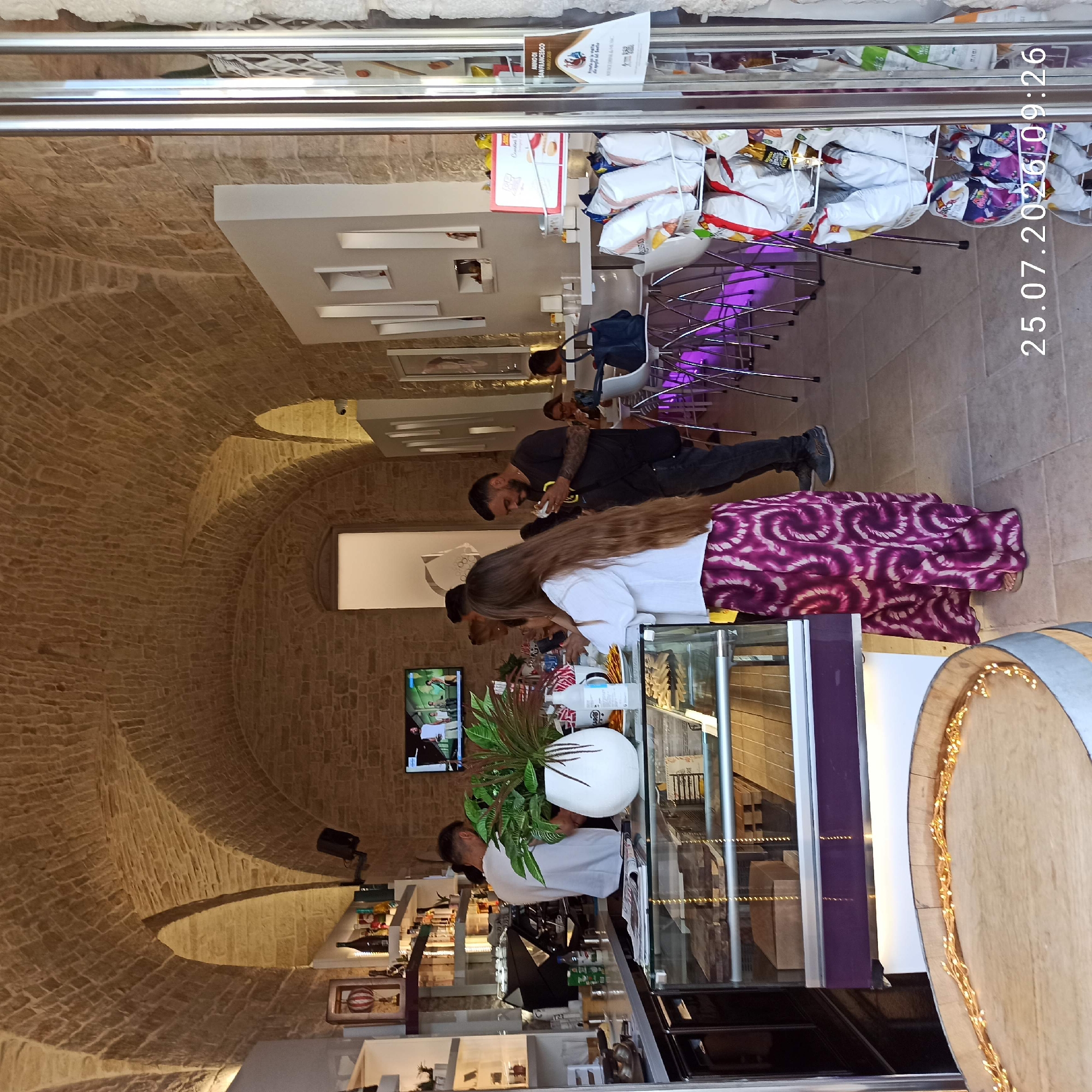 The image depicts the interior of a contemporary cafe or bar with a rustic stone arched ceiling. In the foreground, to the left, is a wooden barrel adorned with fairy lights. A glass display case runs along the counter, filled with pastries. A person in a white shirt and a long, patterned skirt is standing behind the counter, facing away from the camera. To the right of this person, a man in a black t-shirt is visible, holding and eating something. Other patrons are seated at tables in the background. The cafe has modern white shelves and seating, contrasted with the exposed stone walls and ceiling. A television screen displaying a video is mounted on the stone wall. In the far right of the image, a rack of snacks is visible through a doorway or entrance. The timestamp "25.07.2026 09:26" is overlaid on the bottom right corner. This cafe is located in the city of Ruvo di Puglia, Italy, specifically on Via Madonna delle Grazie.