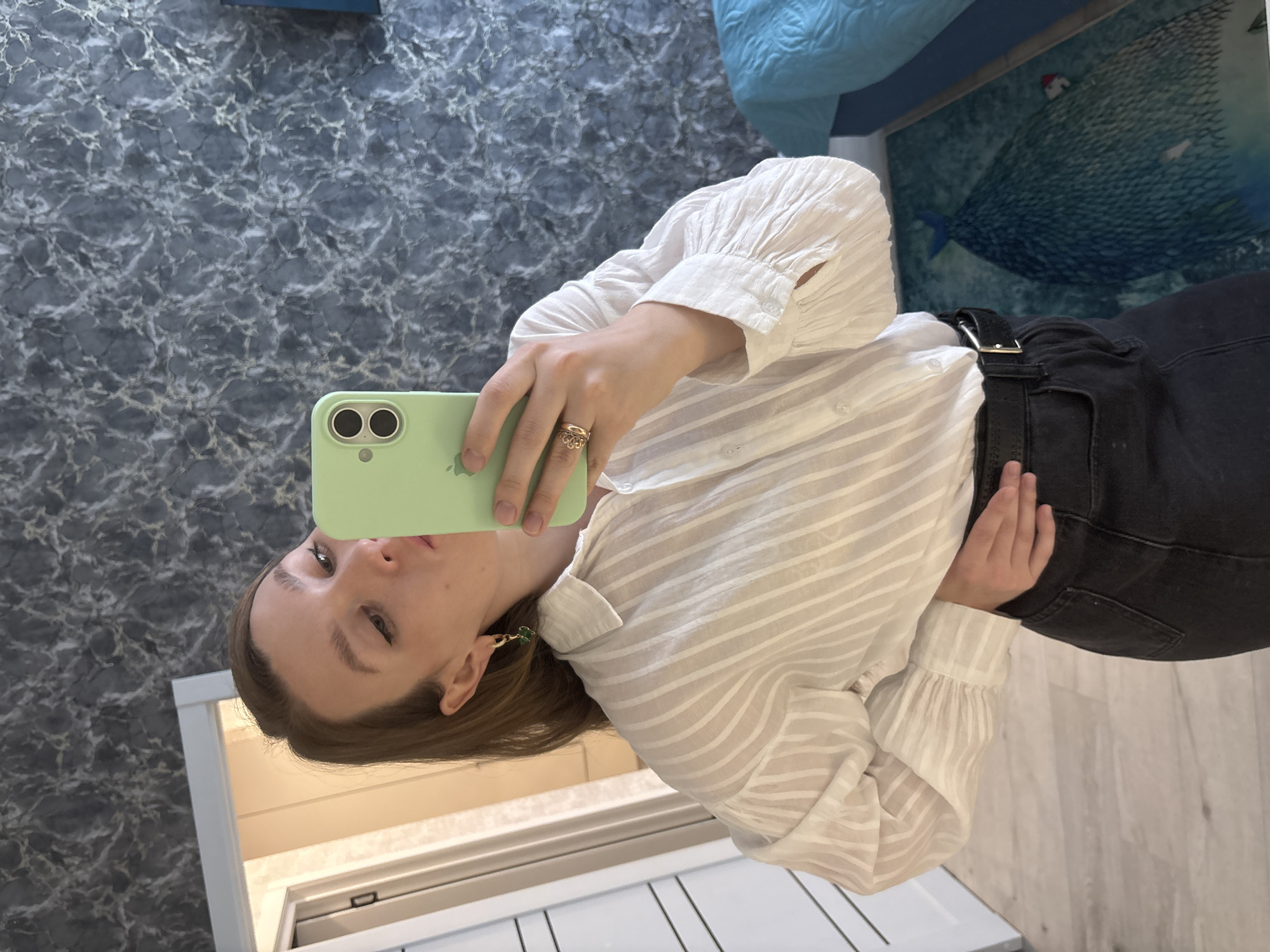 A young woman with shoulder-length brown hair is taking a selfie in a room with a dark, marbled wallpaper. She is wearing a white, semi-sheer, striped button-down shirt with voluminous sleeves and black jeans with a black belt. She has a gold ring on her right ring finger and a gold earring with a green stone in her left ear. She is holding a light green smartphone in her right hand, angled to capture her reflection. Her left hand is resting on her hip. A doorway with white trim is visible on the left side of the frame, and a blue-covered bed is partially visible on the right. The floor is light-colored wood, and a rug with a blue and green abstract design featuring a large, dark, pointed shape is visible in the lower right portion of the image. The room appears to be an indoor residential space in Izhevsk, Russia, given the context provided.
