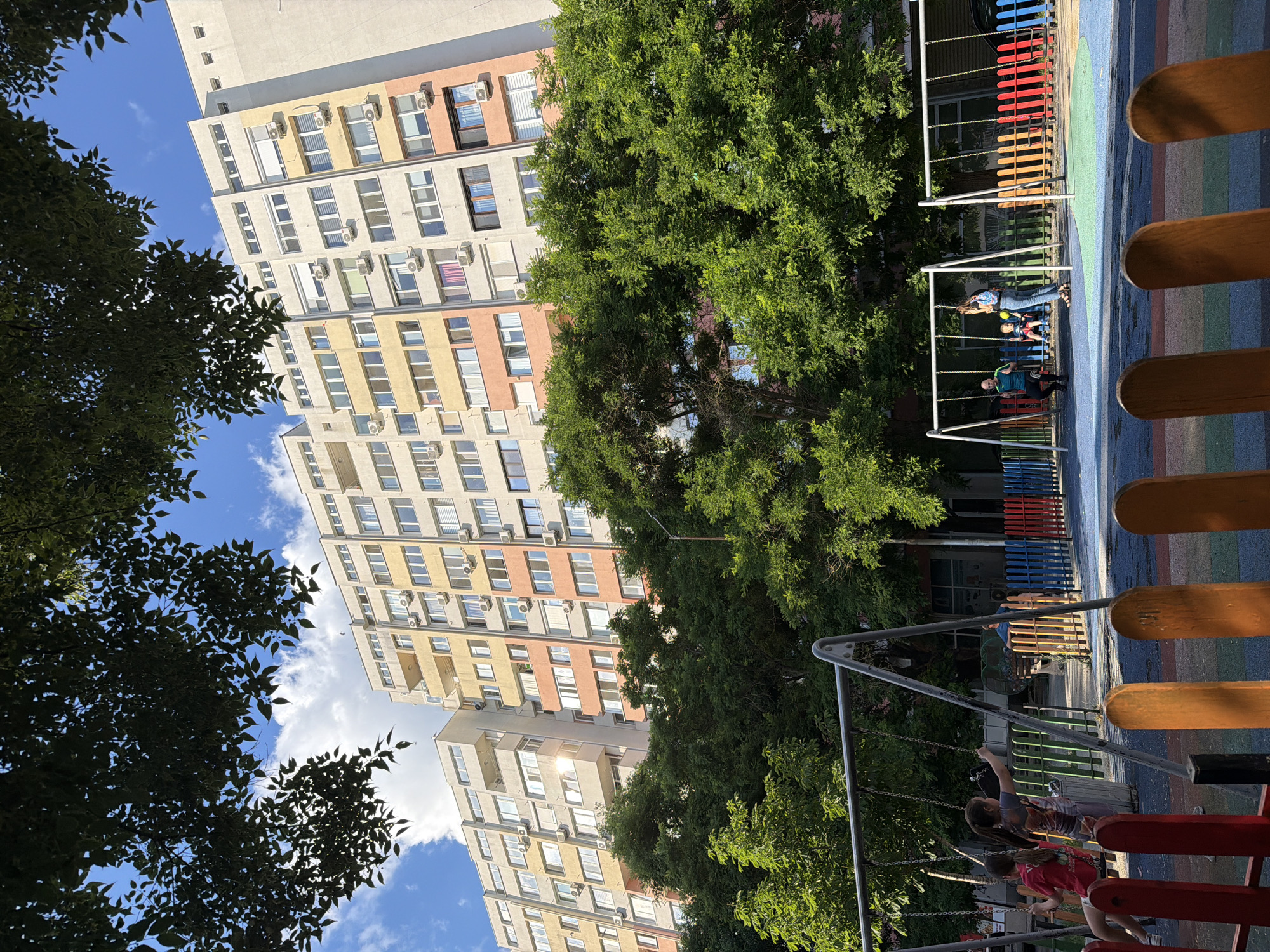 The image shows a low-angle view of a residential building and a playground. The building is tall and multi-story, with a facade featuring alternating sections of light yellow, pink, and beige. Numerous windows and air conditioning units are visible on the building. In the foreground, a playground area is partially visible, with colorful fencing and wooden benches. Two children are on a swing set in the playground. The sun is shining, casting shadows and glints of light on the building, suggesting it is daytime. The sky is blue with scattered clouds. Lush green trees partially obscure the view of the building, framing the scene. The overall atmosphere appears to be a pleasant, sunny day in a residential area with recreational facilities for children.