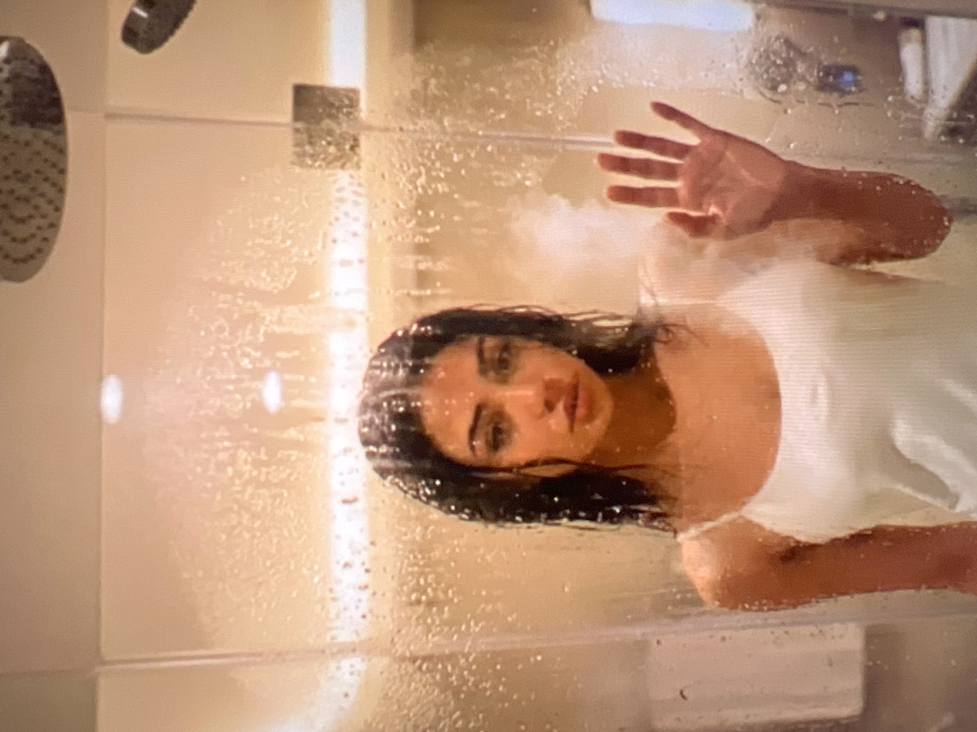 A young woman with dark hair stands behind a glass shower door that is covered in water droplets, obscuring her body partially. She is wearing a white slip. Her left hand is pressed against the glass, palm facing outwards. The background appears to be a tiled wall and a shower head. The lighting is warm and diffused. There is no indication of the city of Athens, Greece.
