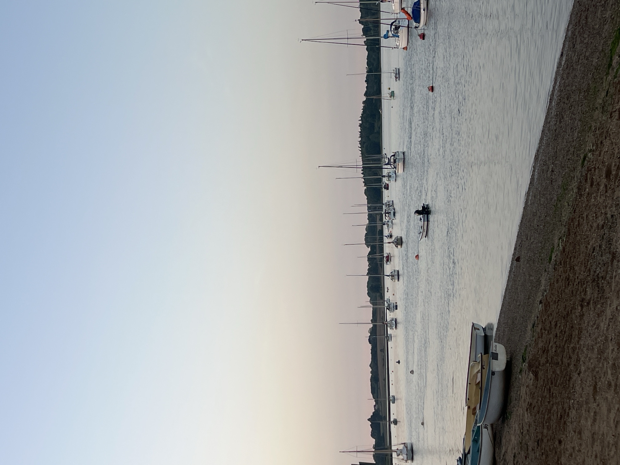 The image depicts a body of water, possibly an estuary or harbor, in East Suffolk, United Kingdom, at dusk or dawn. A line of sailboats, recognizable by their masts, are moored or anchored in the water. Along the right edge of the image is a shoreline with beach. A small motorboat with one or two people inside is visible on the water. The sky is a gradient of light blue to light pink.